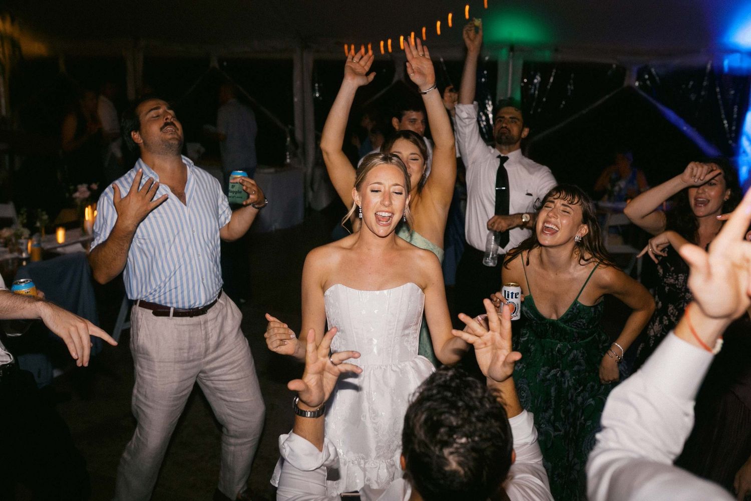 People dancing and celebrating around a bride and groom at a lively wedding reception with colorful lights in the background.