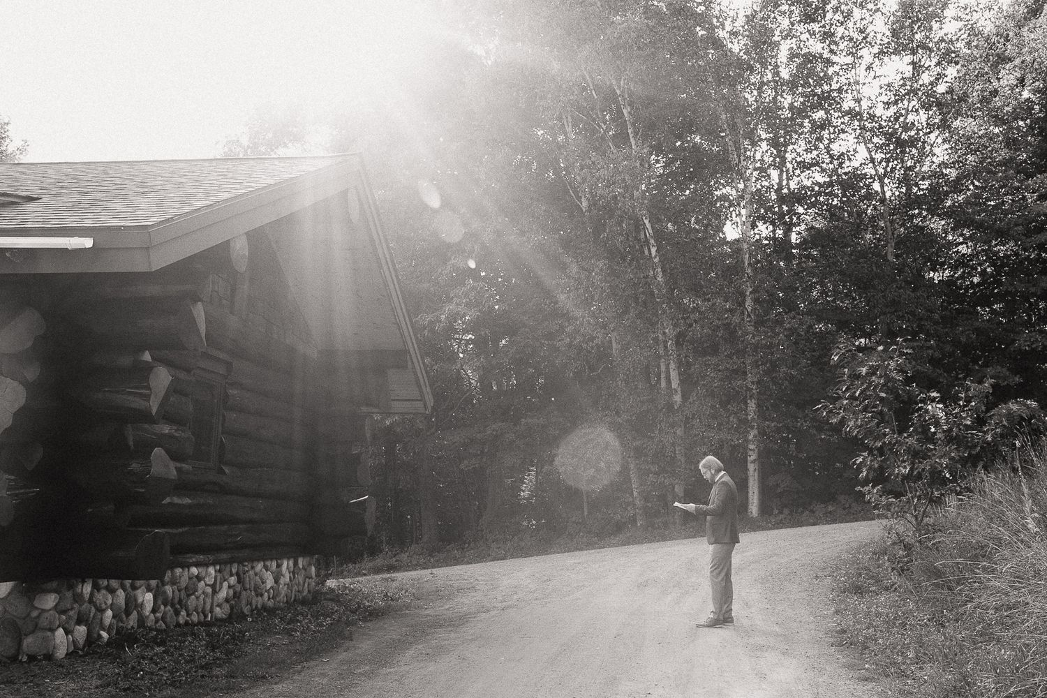 Black and white photo of a person reading in golden sunlight outside a rustic log cabin.