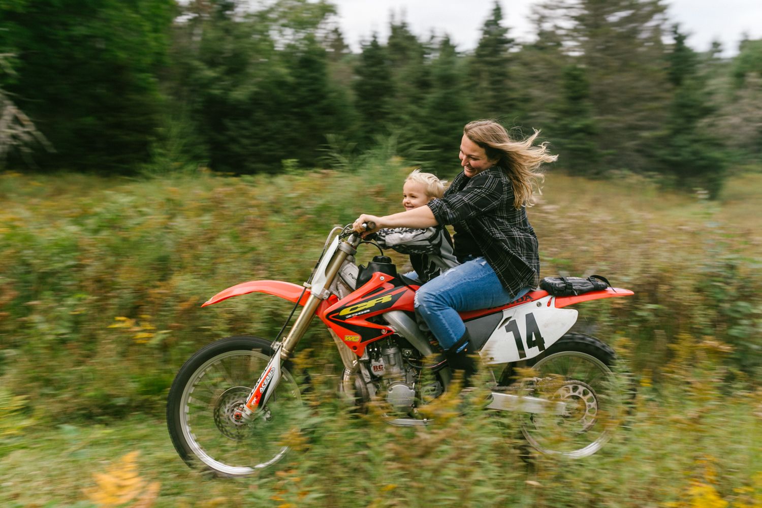 A woman and child ride a red dirt bike number 14 through a field at high speed.