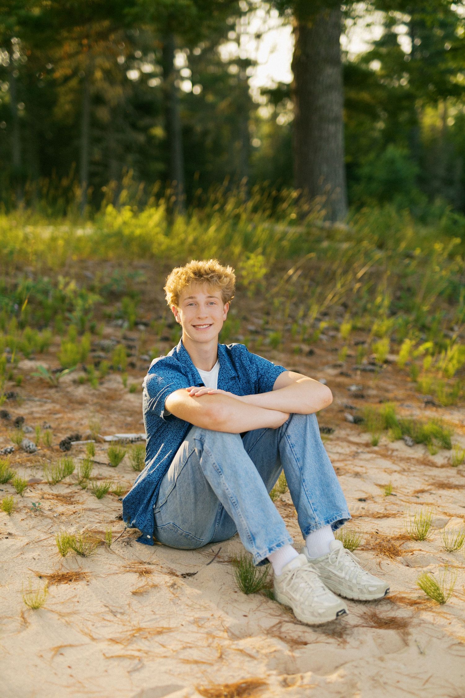 Young man with curly hair sitting on sandy ground in denim outfit, smiling in golden hour forest light.