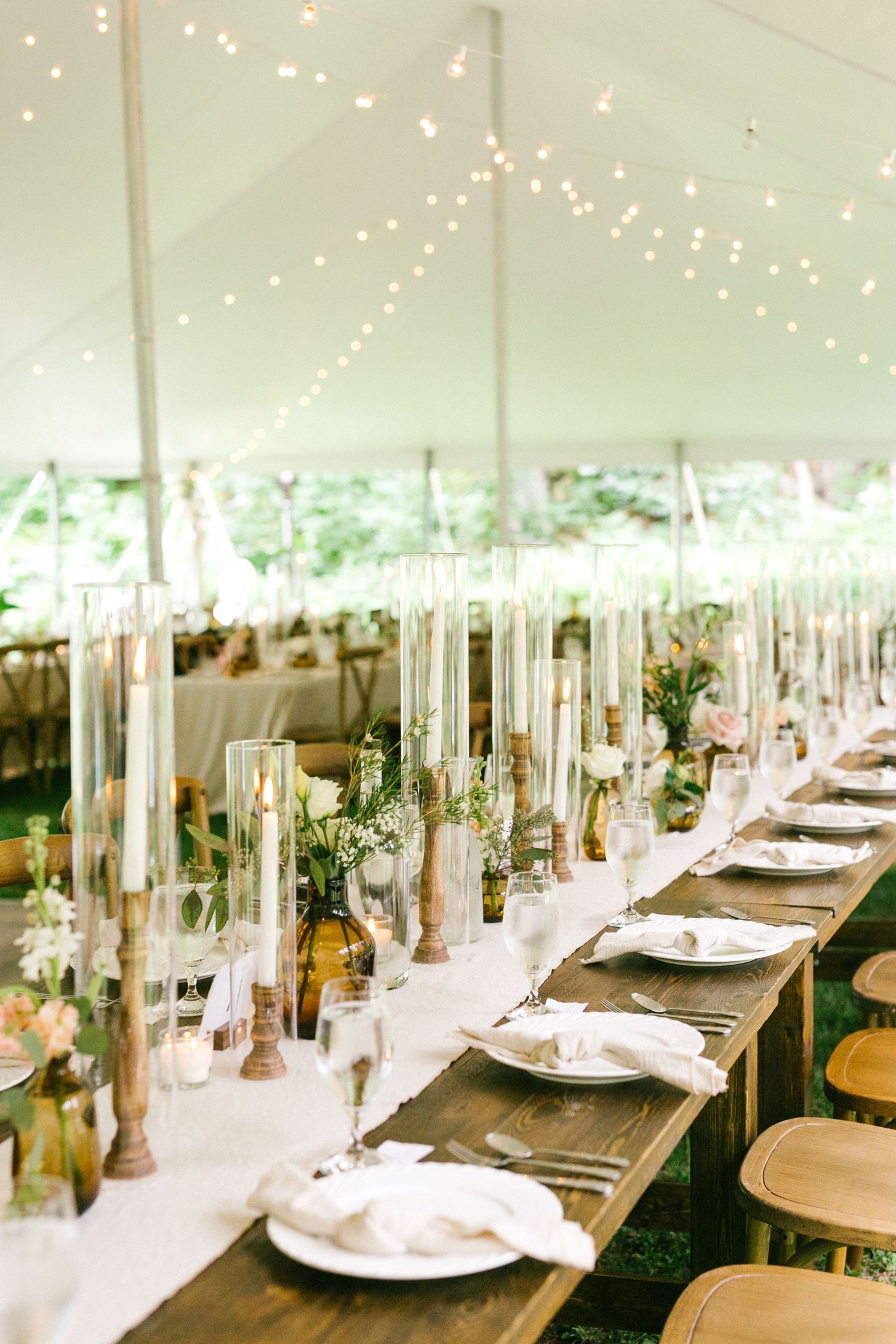 Elegant wedding reception tables set under a white tent with string lights and tall glass centerpieces creating a romantic atmosphere.
