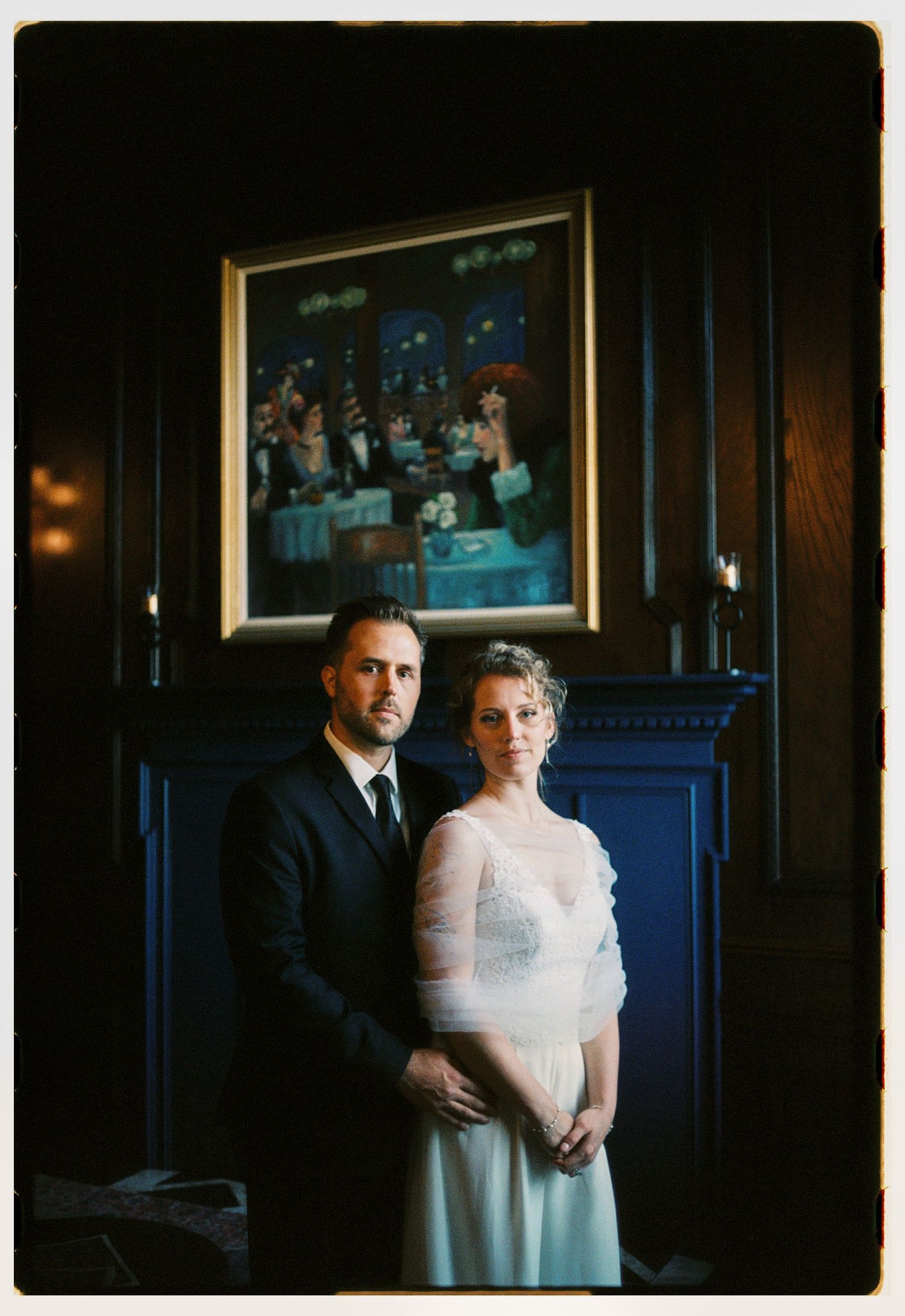 Elegant wedding couple posing before a framed painting in a dark wood-paneled room, bride in white gown, groom in dark suit.