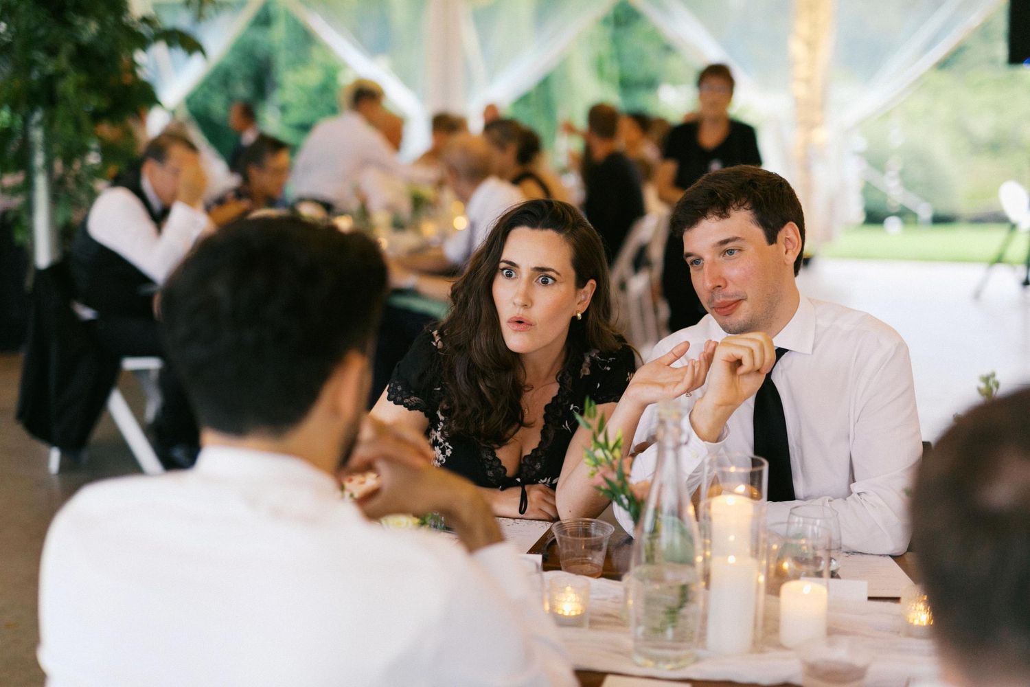 Wedding guests enjoying conversation and drinks at elegant reception tables with white linens and floral centerpieces.