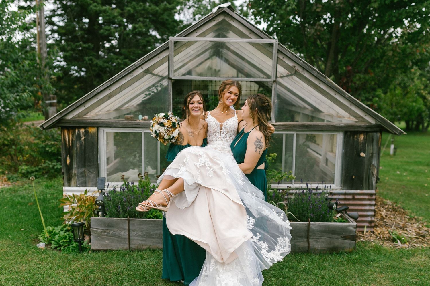 Three people in formal attire pose in front of a rustic greenhouse with glass panels and wooden frame in a garden setting.