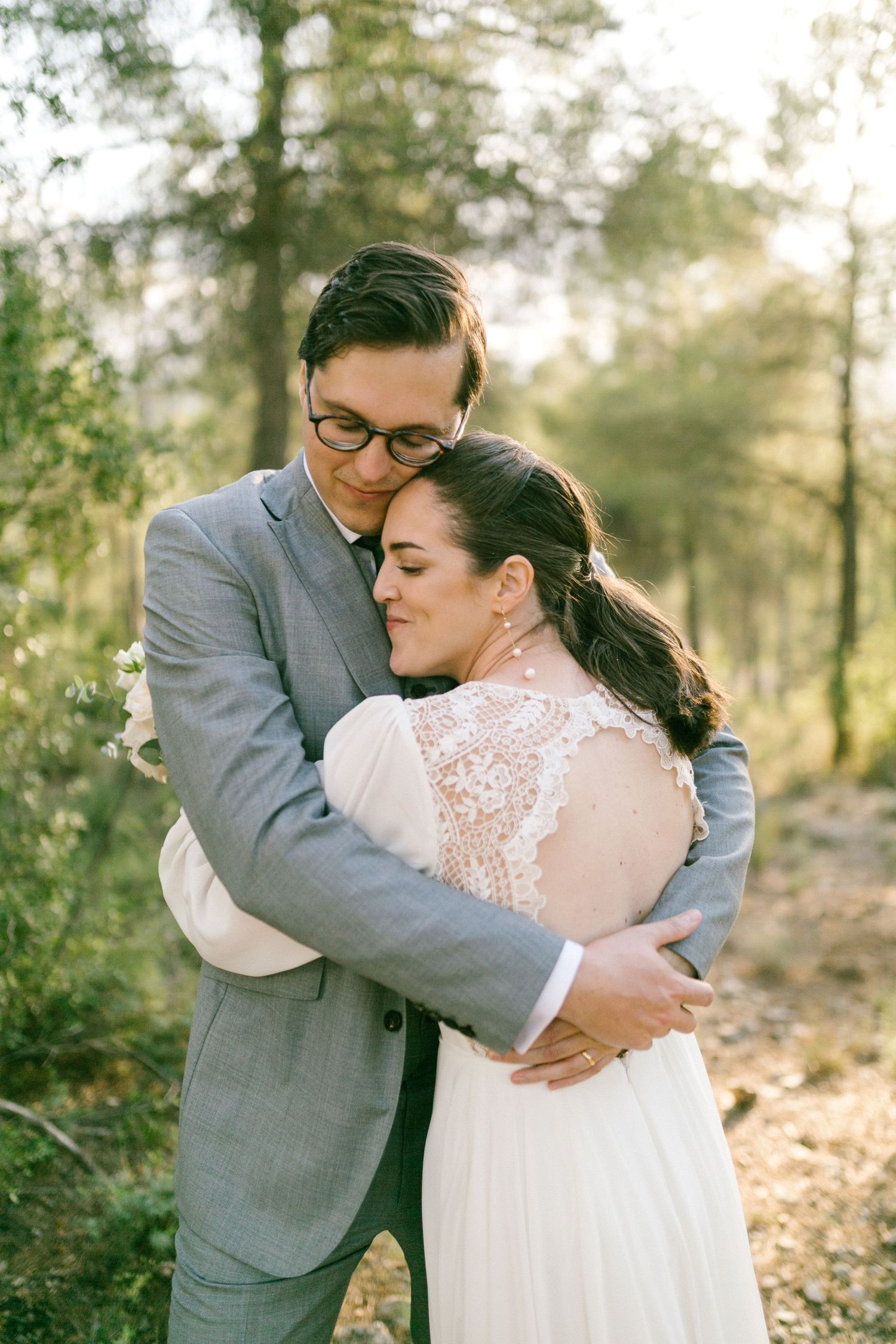 A tender embrace between newlyweds in a sunlit forest, with the bride wearing a delicate lace dress and groom in gray suit.