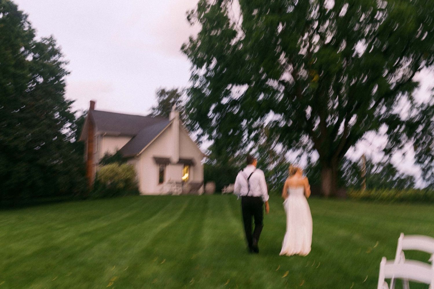 A couple stands on a green lawn outside a rustic house during a wedding ceremony on an overcast day.