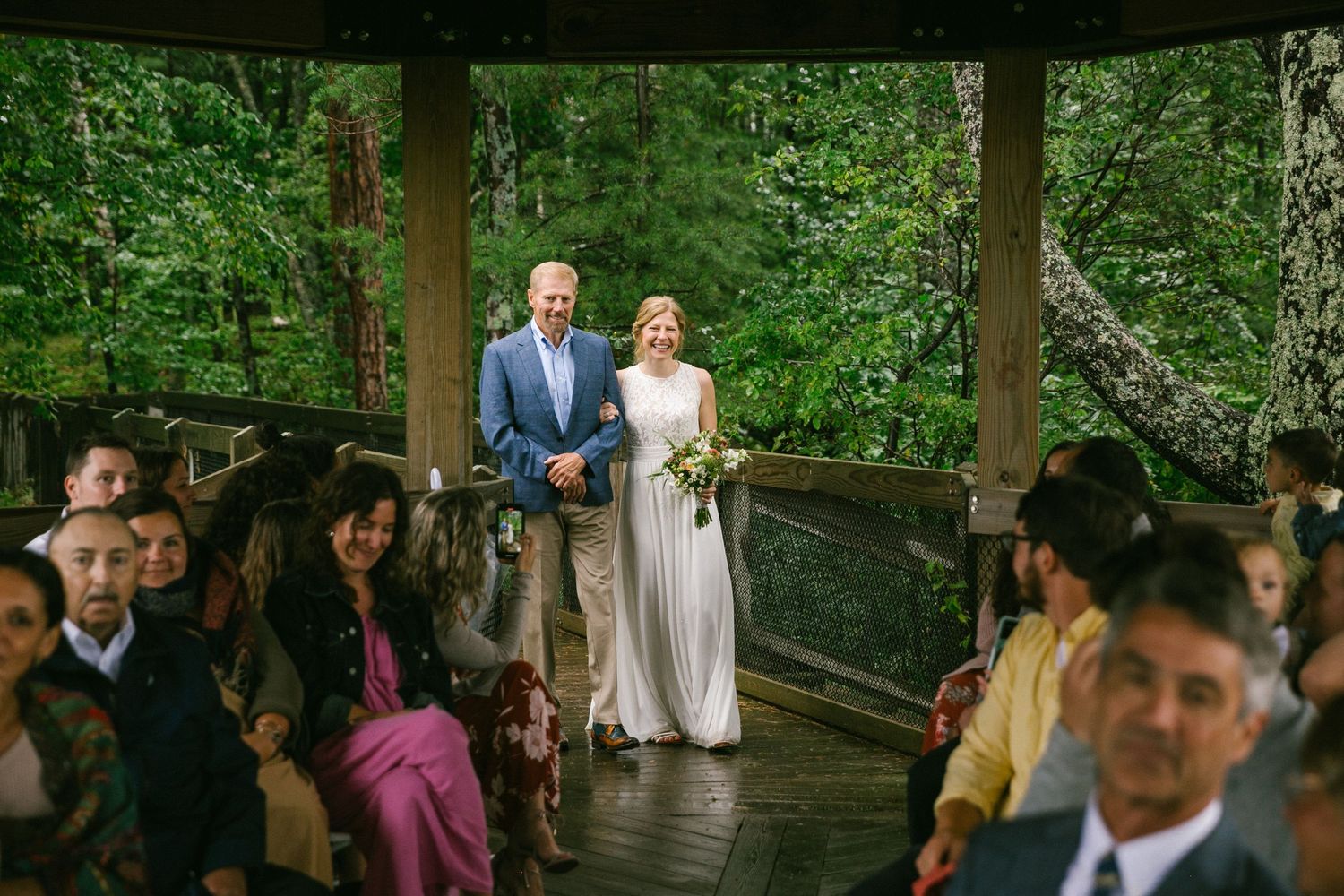 Wedding ceremony taking place in a rustic wooden gazebo surrounded by lush greenery with guests seated.