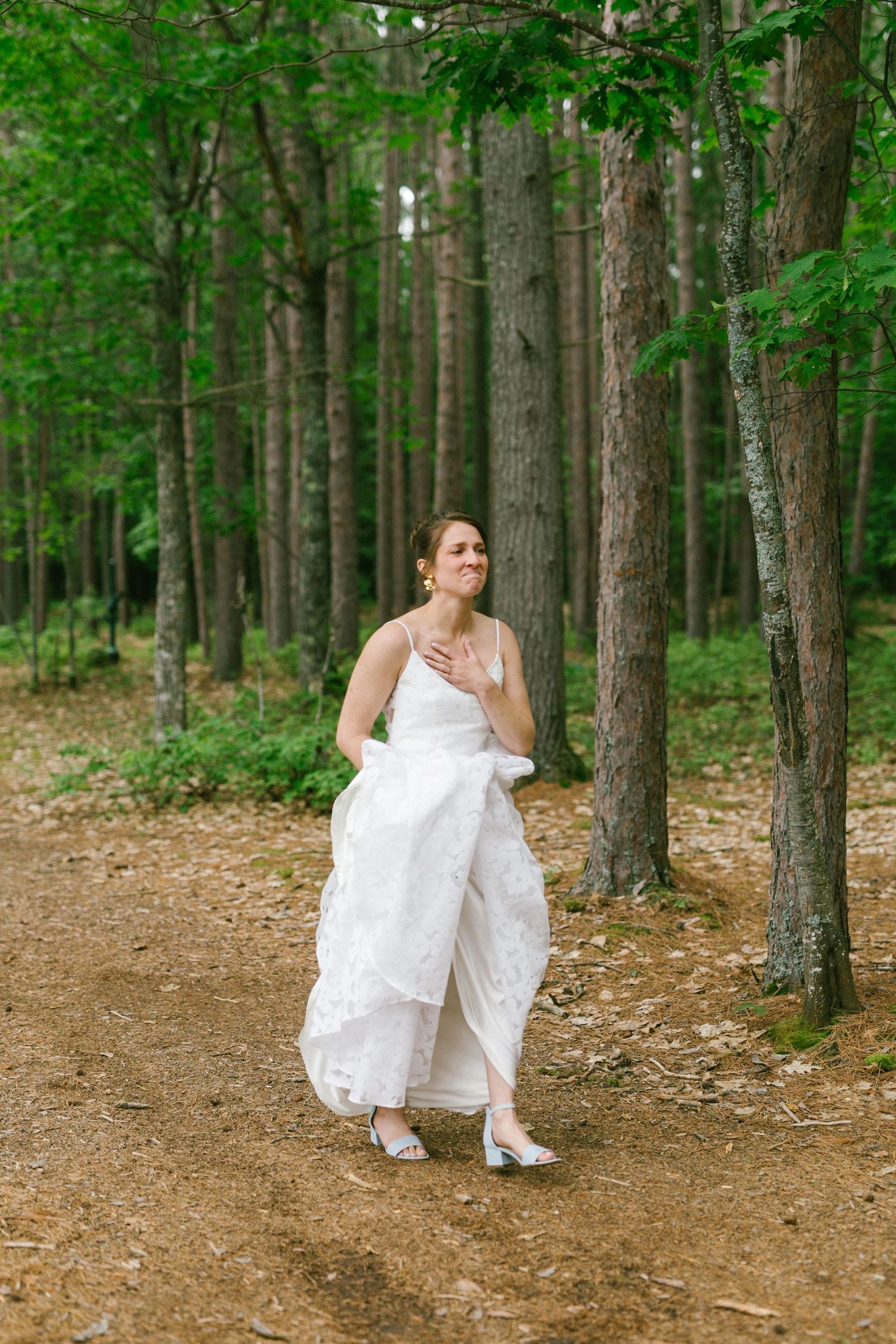 A person in a flowing white dress walks along a dirt path through a lush green forest on a summer day.