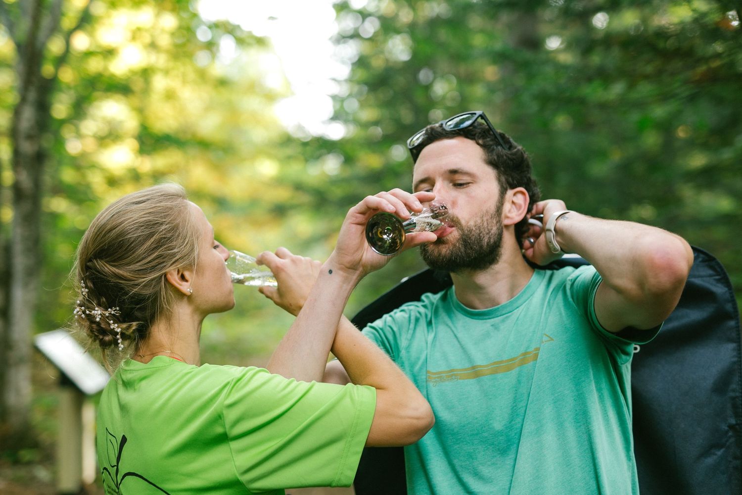 Two people in bright green and turquoise shirts take a drink outdoors in a sunlit forest setting.