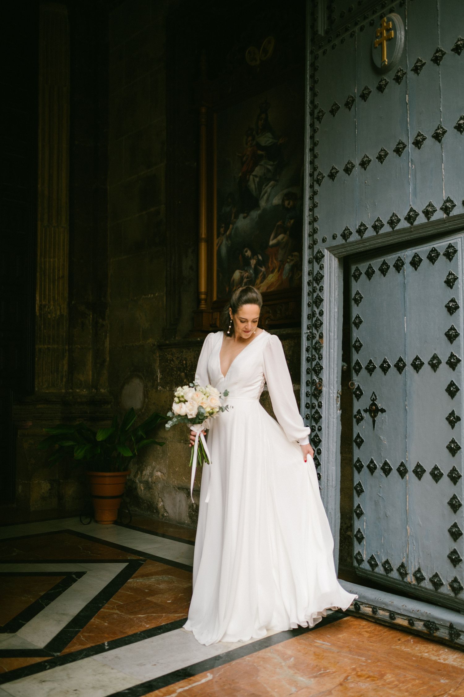 Bride in long-sleeve white wedding gown poses elegantly by ornate metal door in dramatic lighting in a Spanish Cathedral.