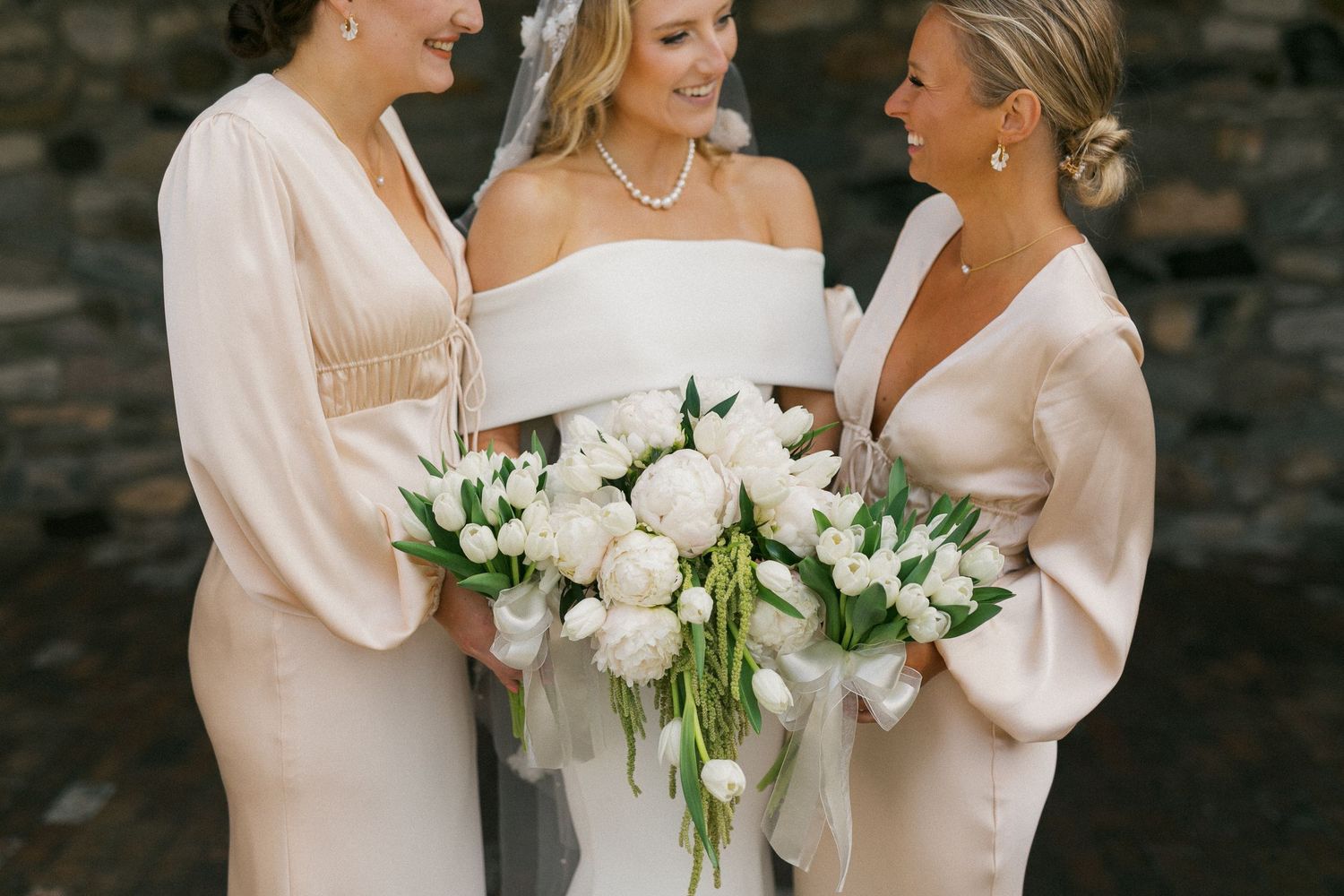 Three women in blush-colored dresses hold white floral bouquets at a wedding.