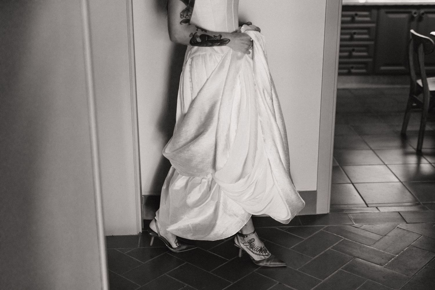 Black and white photo of a bride in a flowing white wedding gown and heels standing in a doorway.