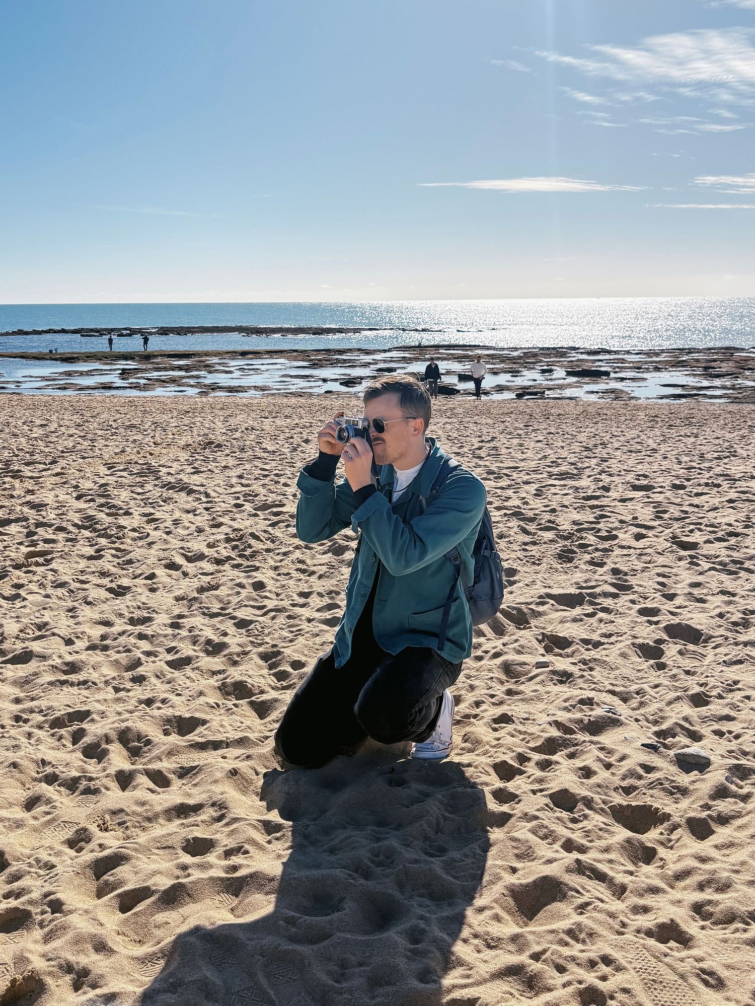 Dylan of Miles north Creative . Co kneeling on sandy beach with camera, ocean horizon and rocky shoreline visible under blue sky.