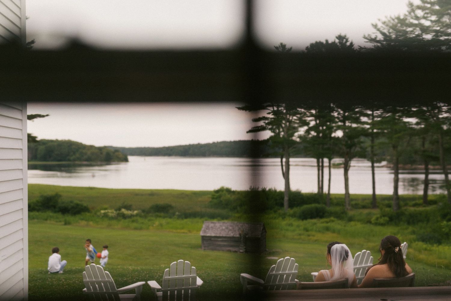People relaxing in Adirondack chairs overlooking a serene lake surrounded by lush green trees and lawn.