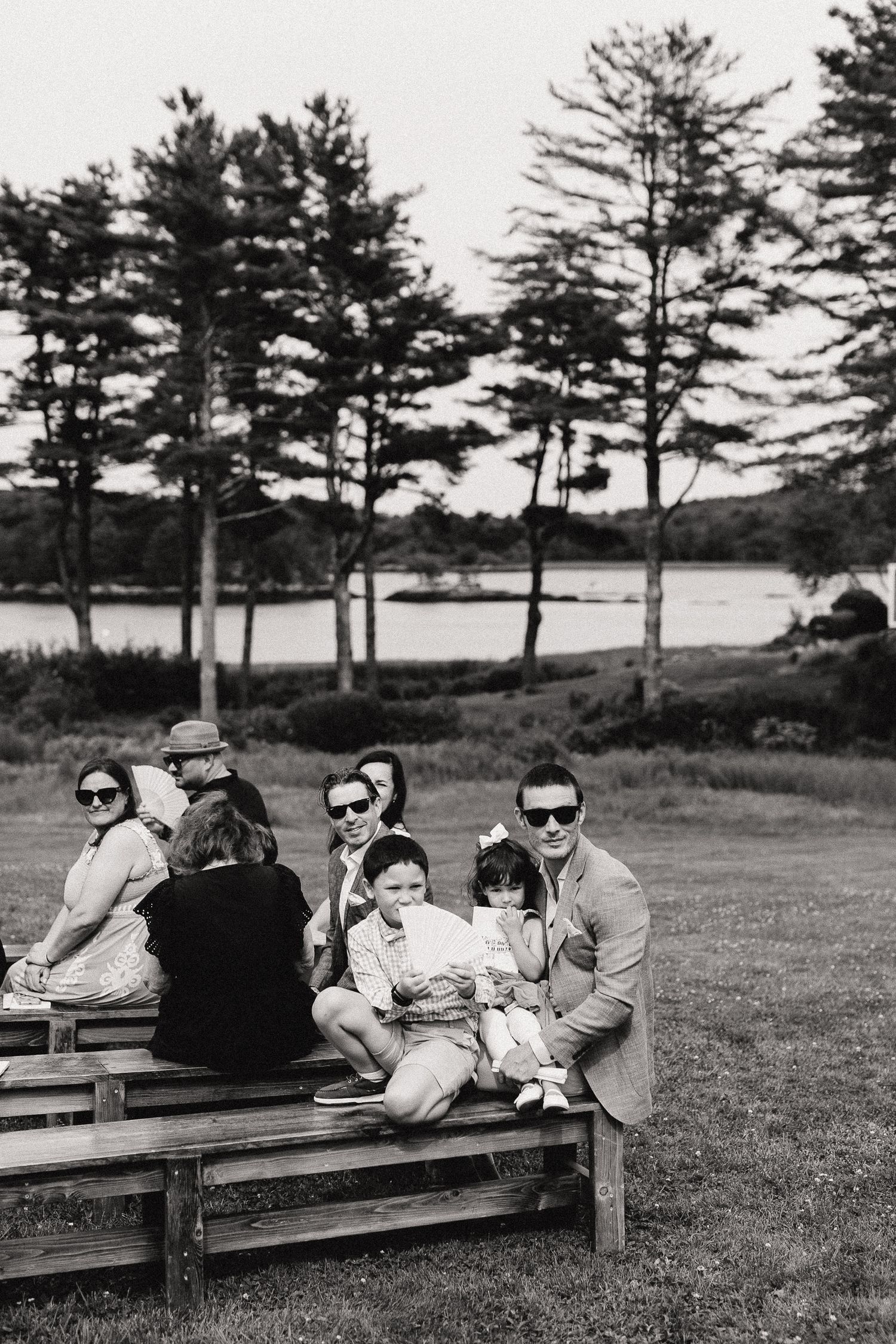 Black and white photo of a family with children sitting on wooden benches outdoors near a lake surrounded by tall pine trees.
