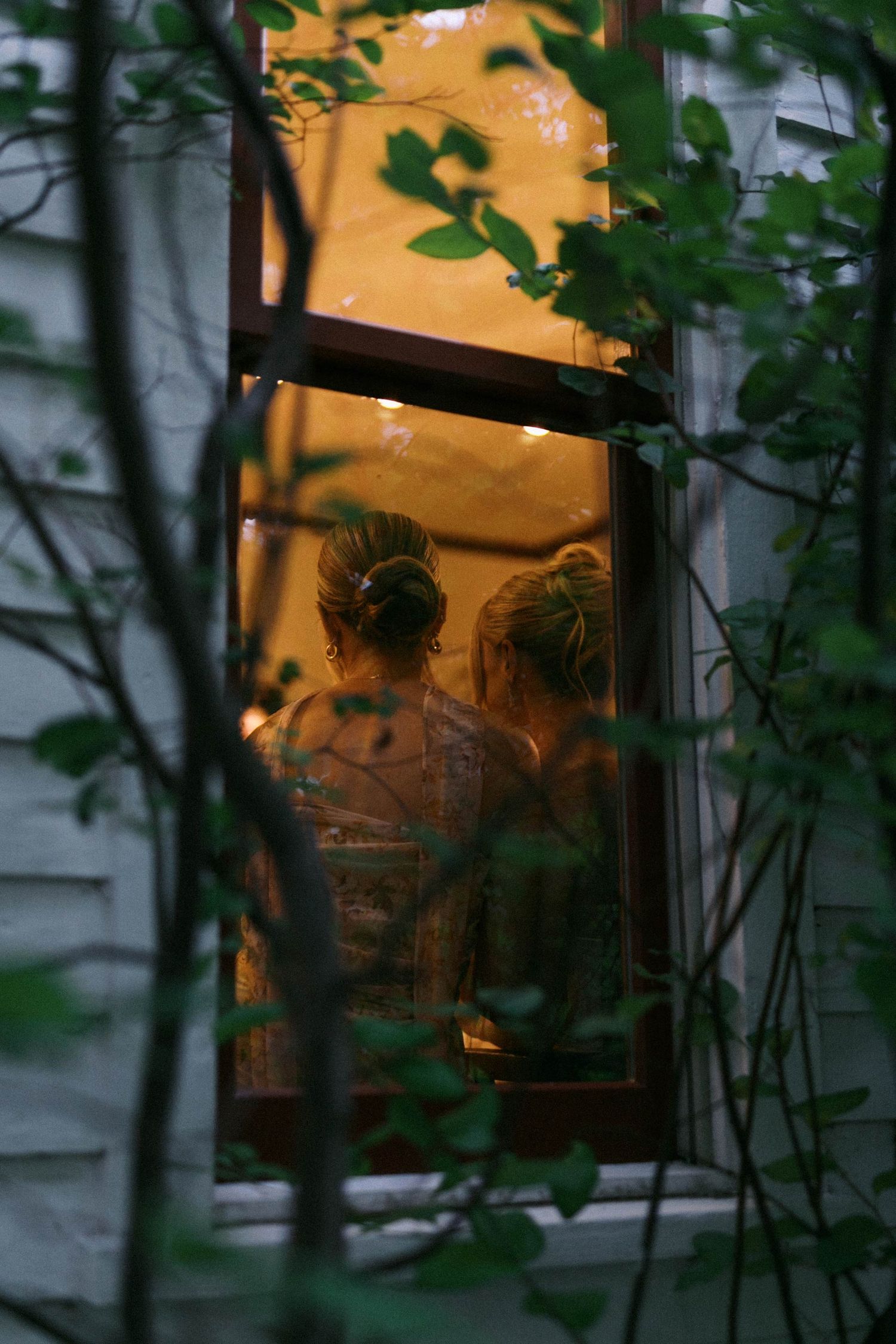 A dimly lit window view through foliage shows a warm orange glow from inside a building at dusk.