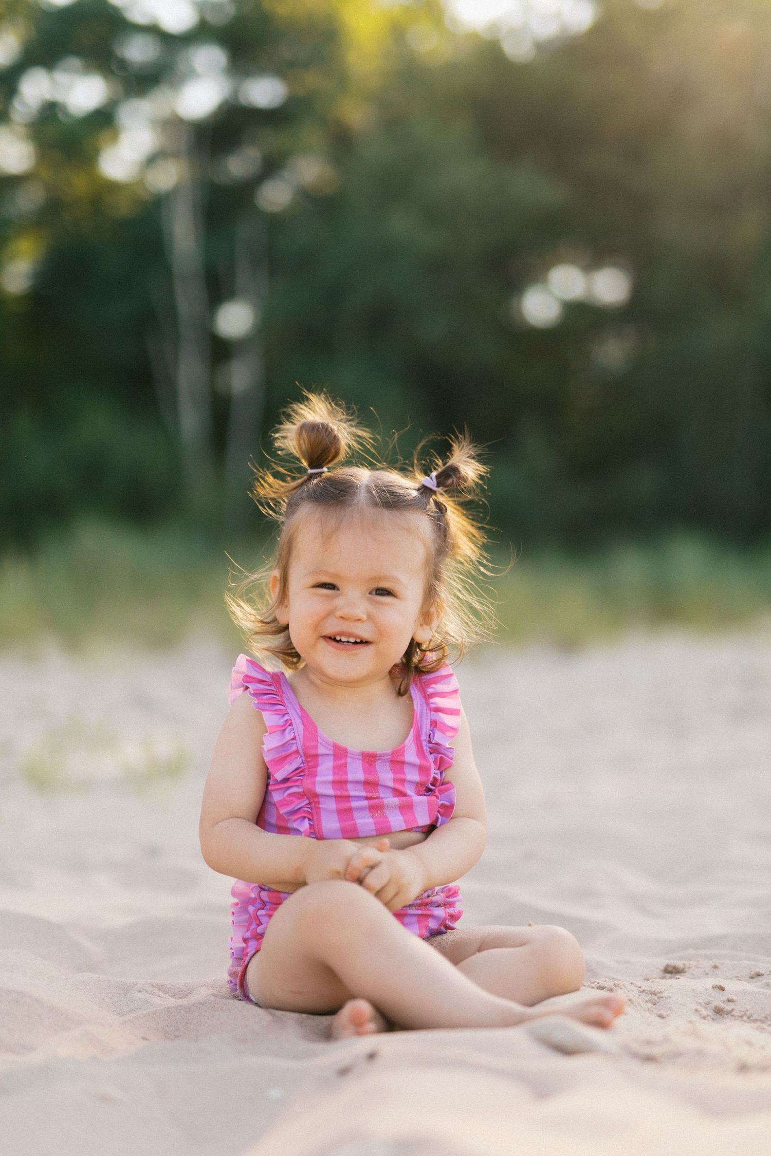 Adorable toddler girl in pink swimsuit sitting on sandy beach smiling with pigtails.