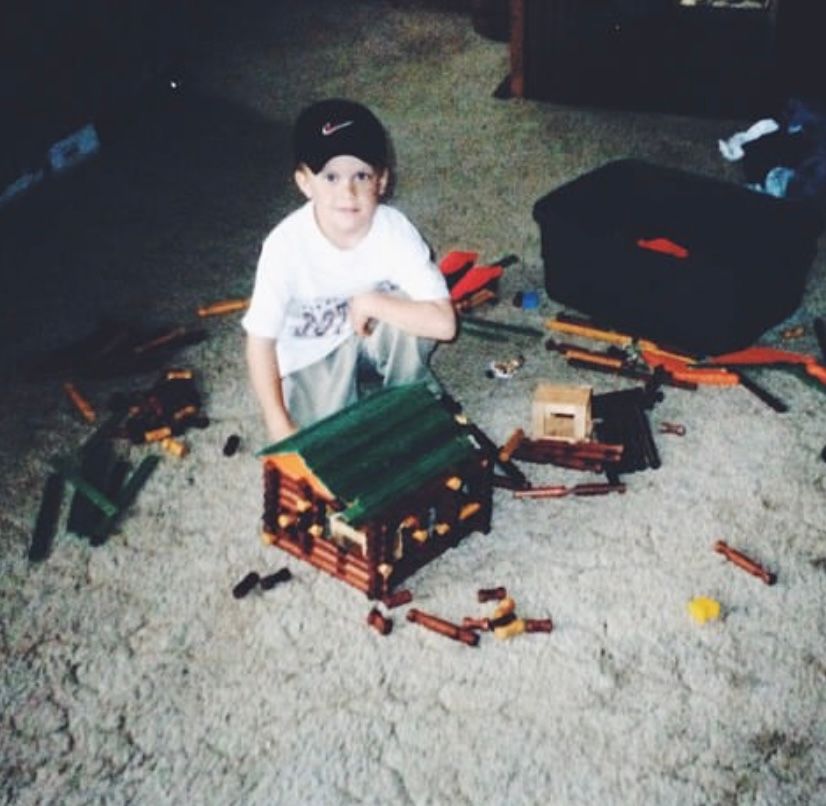 Young boy in Nike cap posing with K'Nex log cabin construction on carpet floor.