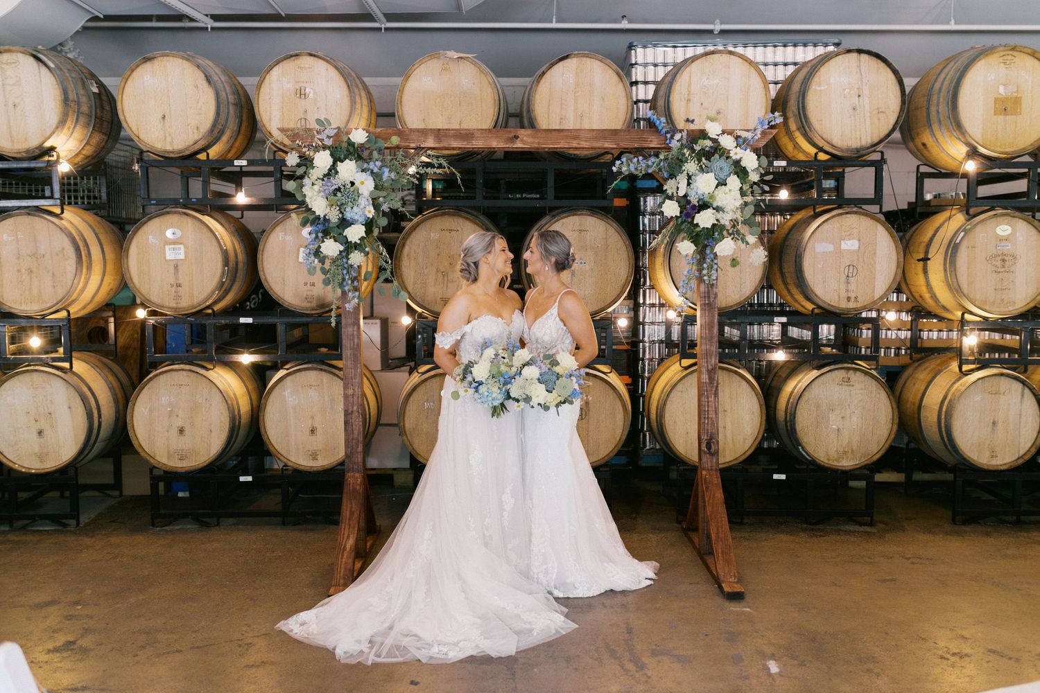 A romantic wedding scene with two people in white dresses seated in front of wooden wine barrels and floral arrangements.
