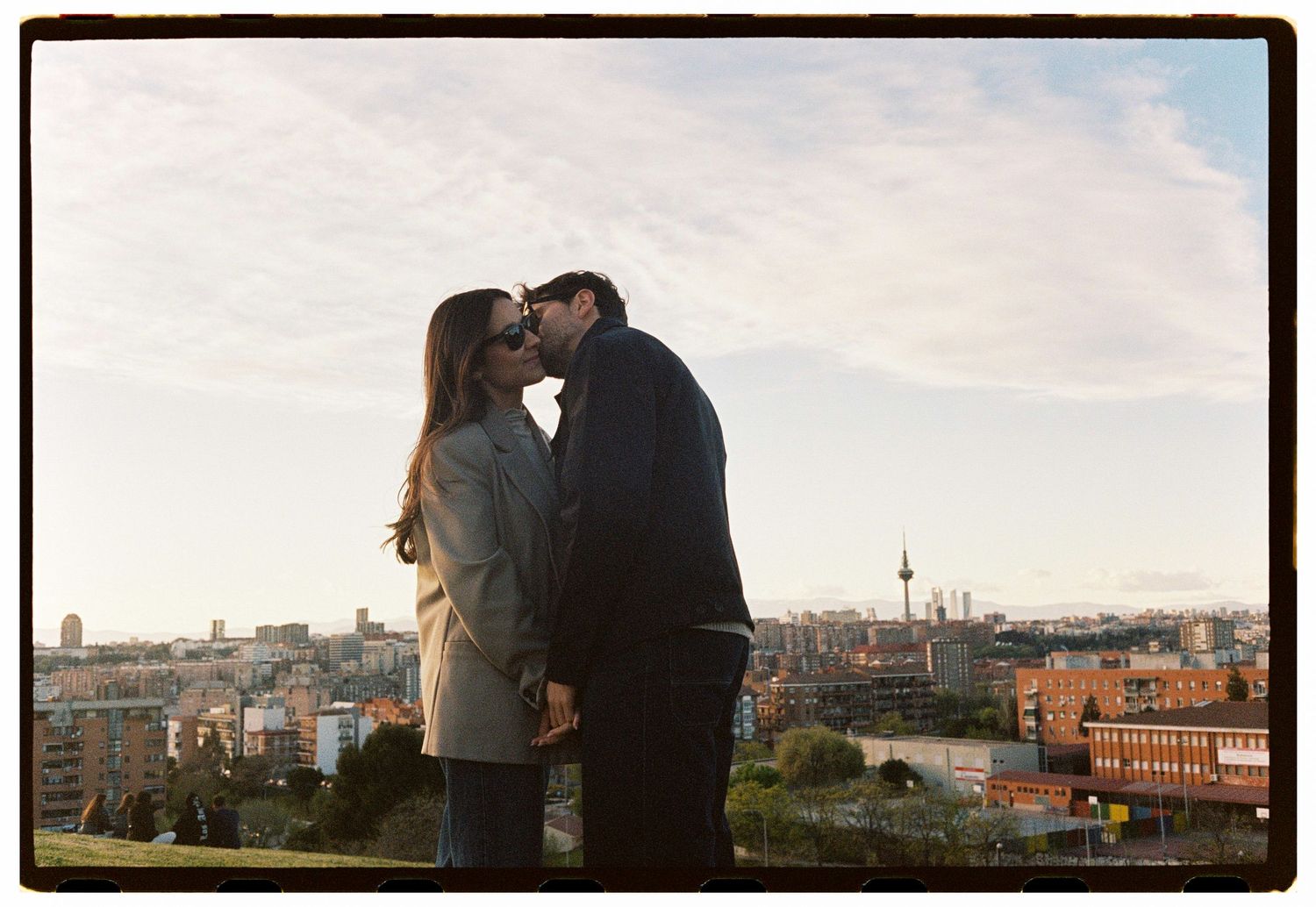 Romantic couple kissing at sunset with a city skyline and TV tower visible behind them.