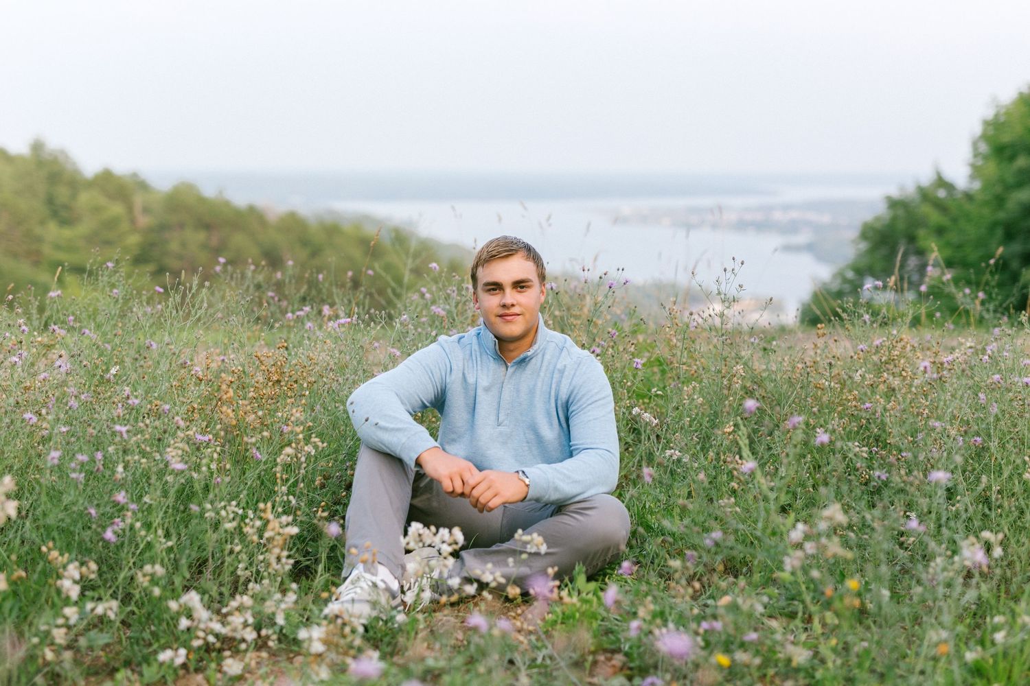 Young man sits relaxed in a wildflower meadow wearing a light blue hoodie with a scenic water view behind him.