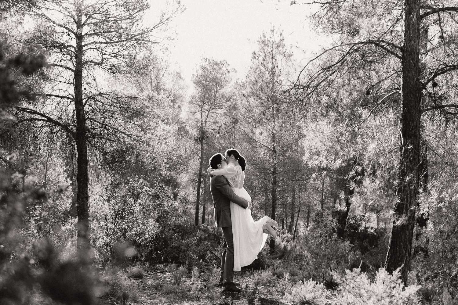 Black and white photo of a couple embracing in a forest, the groom lifting his bride among tall pine trees.