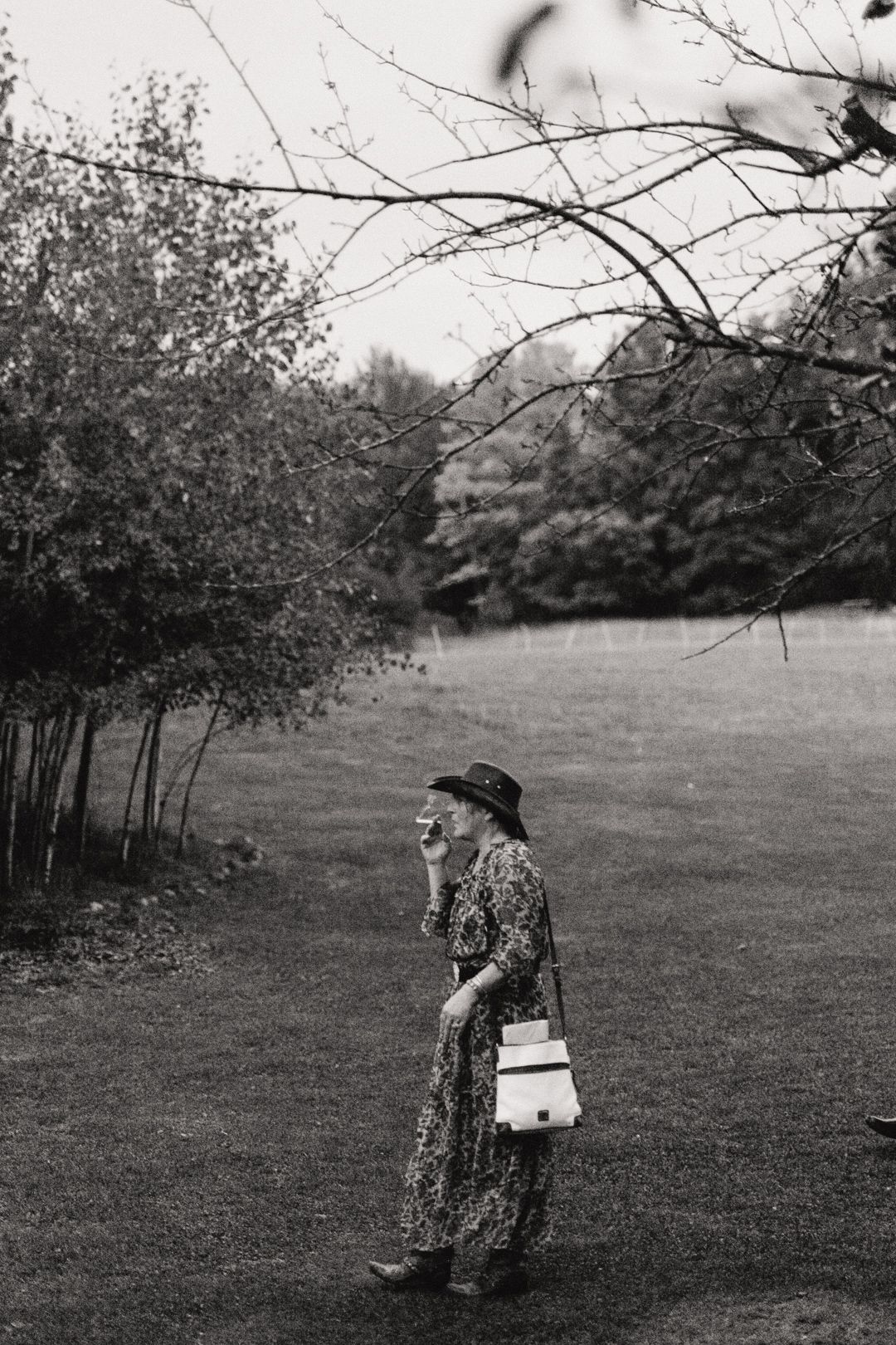 Woman in floral dress and wide-brim hat standing in lush garden, black and white photography.