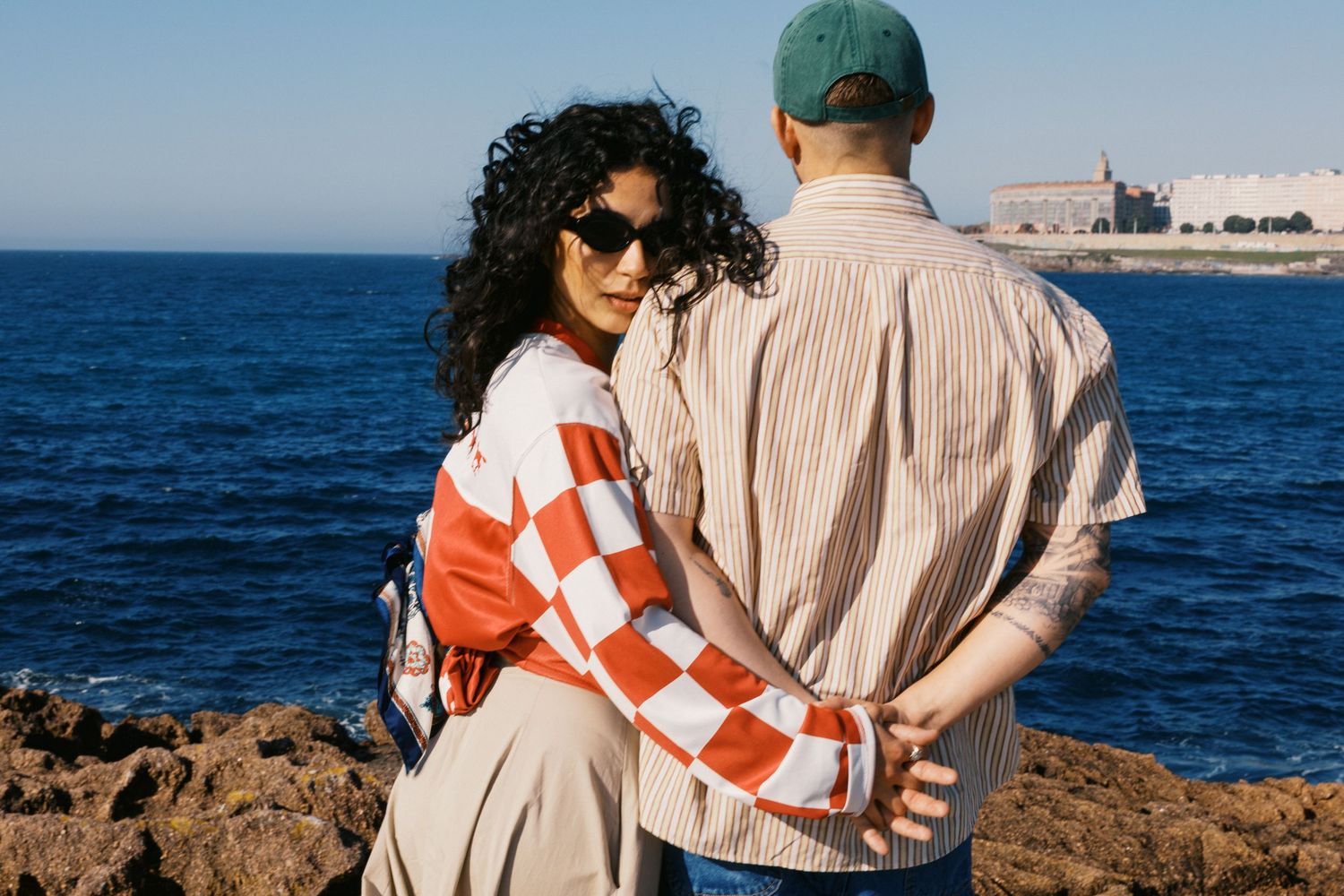 Couple embracing on rocky coastline with blue ocean and historic building in background.