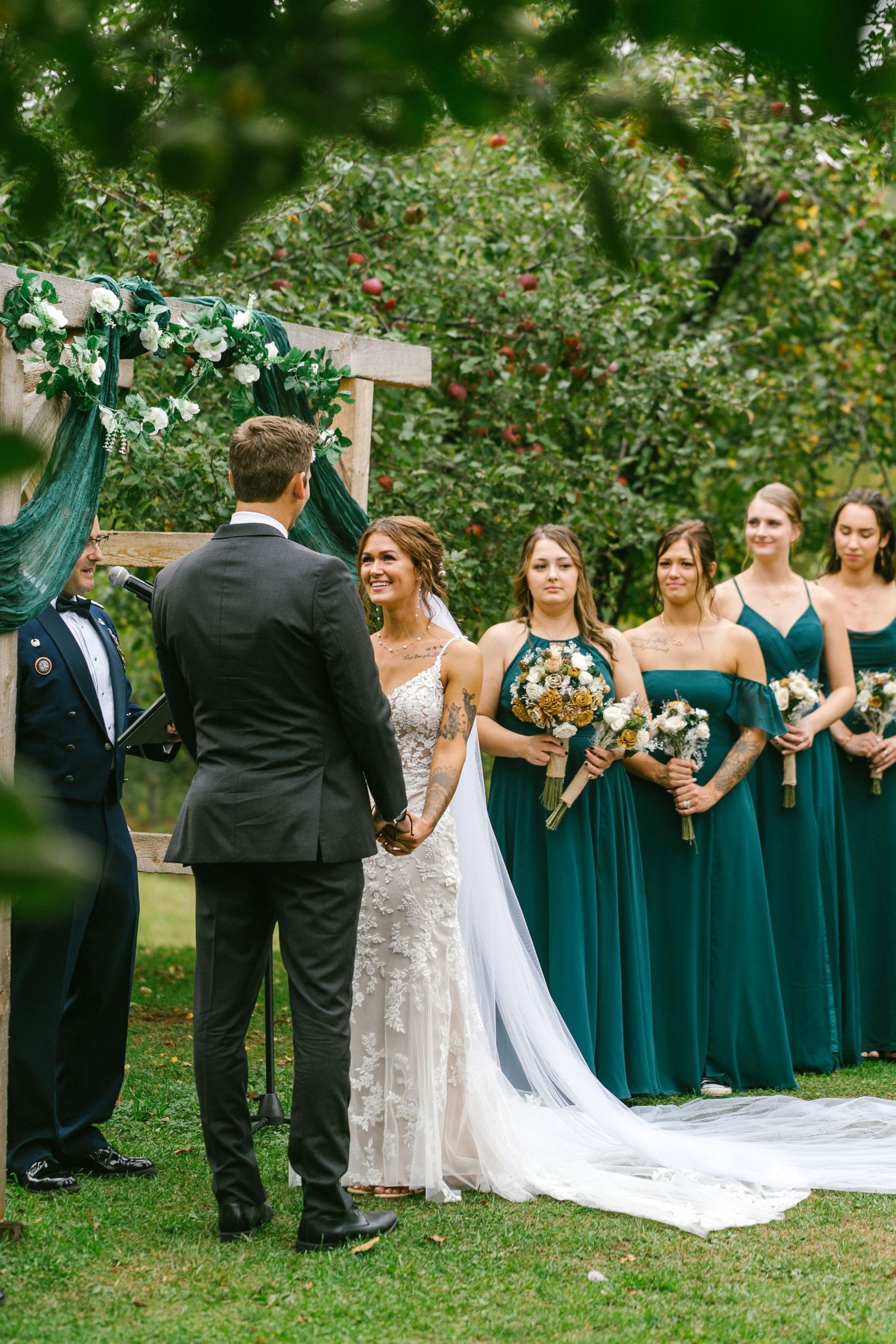Wedding ceremony under a decorated wooden arch with bridesmaids in teal dresses holding bouquets.