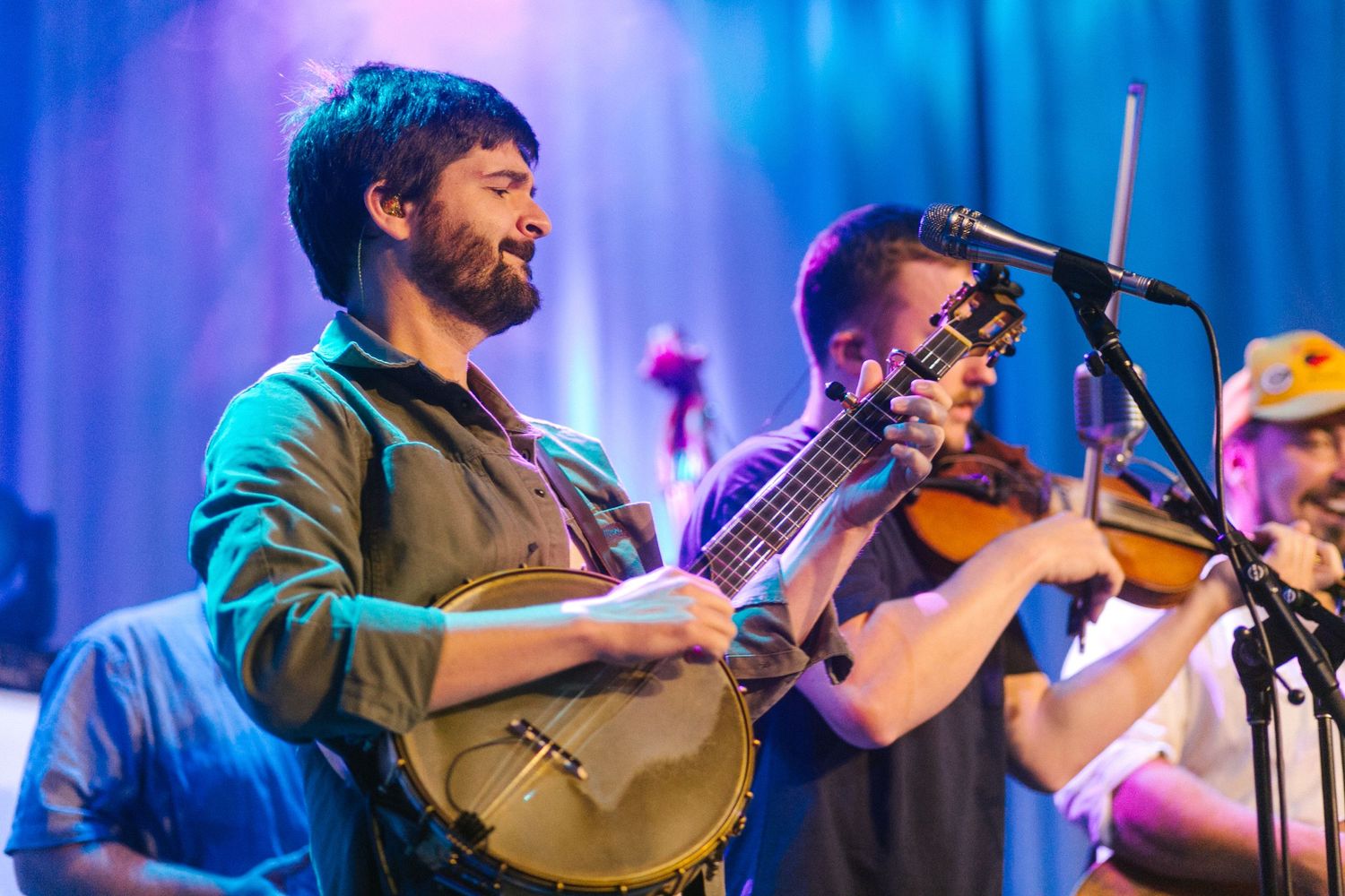Musicians performing on stage with banjo and violin under purple and blue stage lighting.