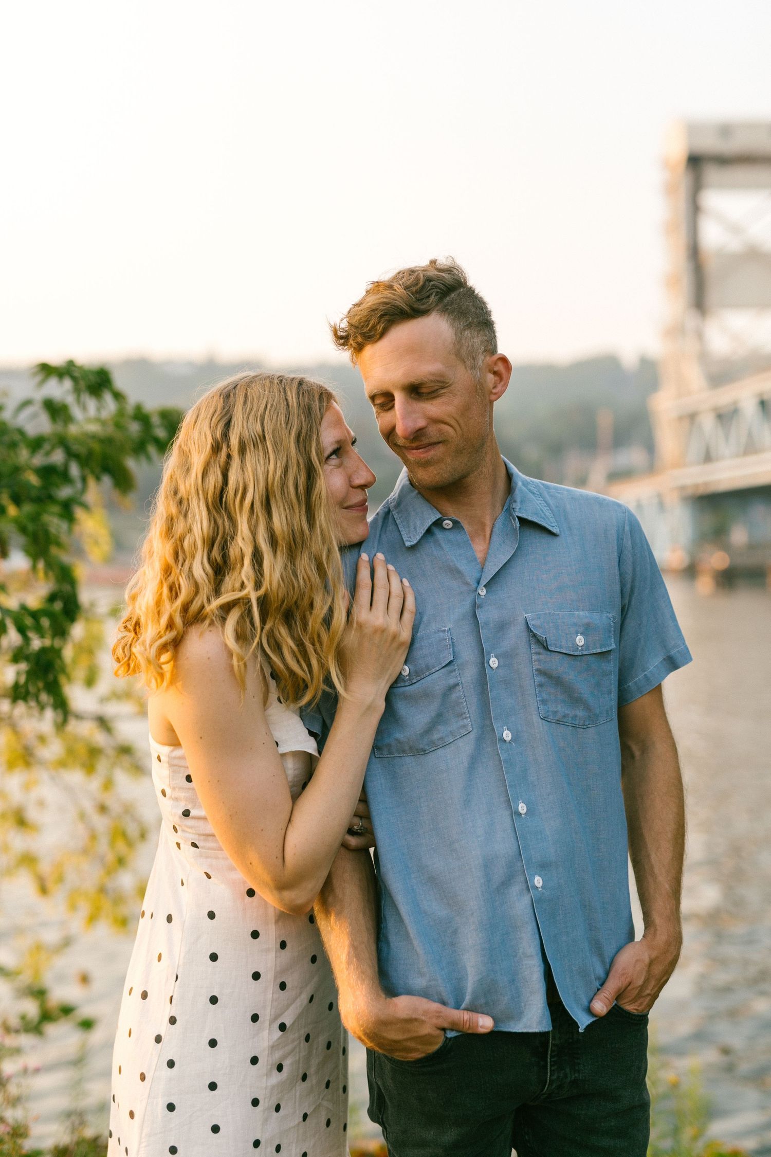 A romantic couple poses together by a waterfront bridge during sunset.