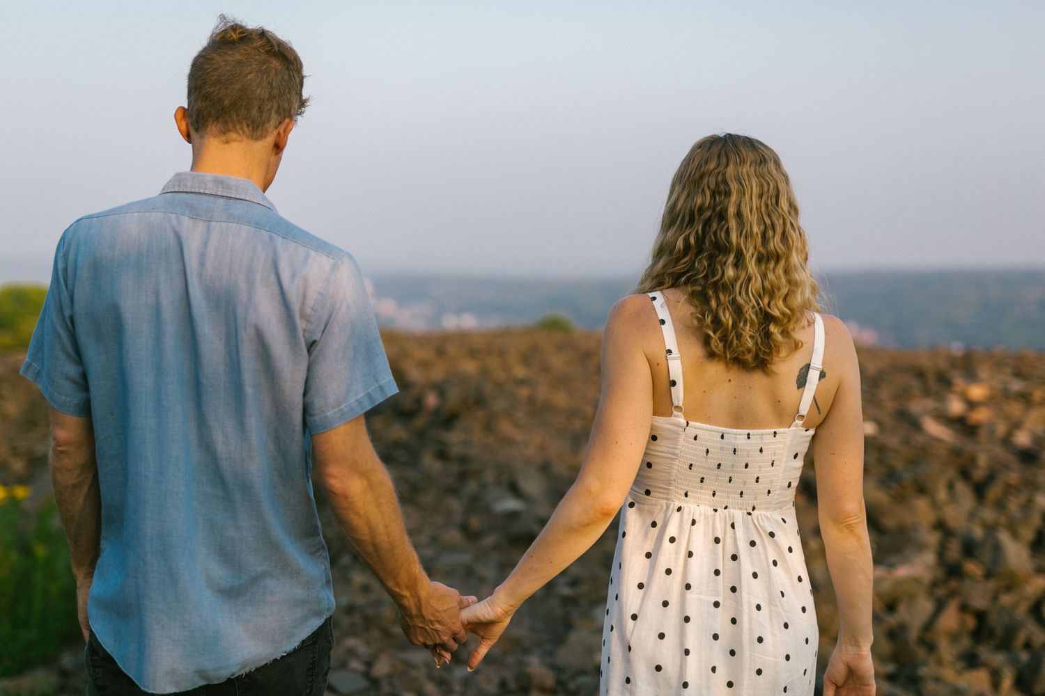 A couple in casual attire holds hands while walking along a rocky overlook at sunset.