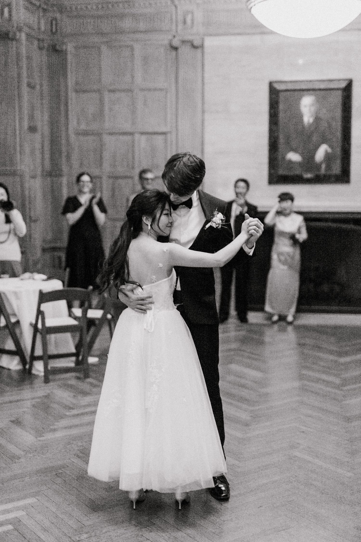 Black and white photo of bride and groom sharing first dance at elegant wedding reception with guests watching.