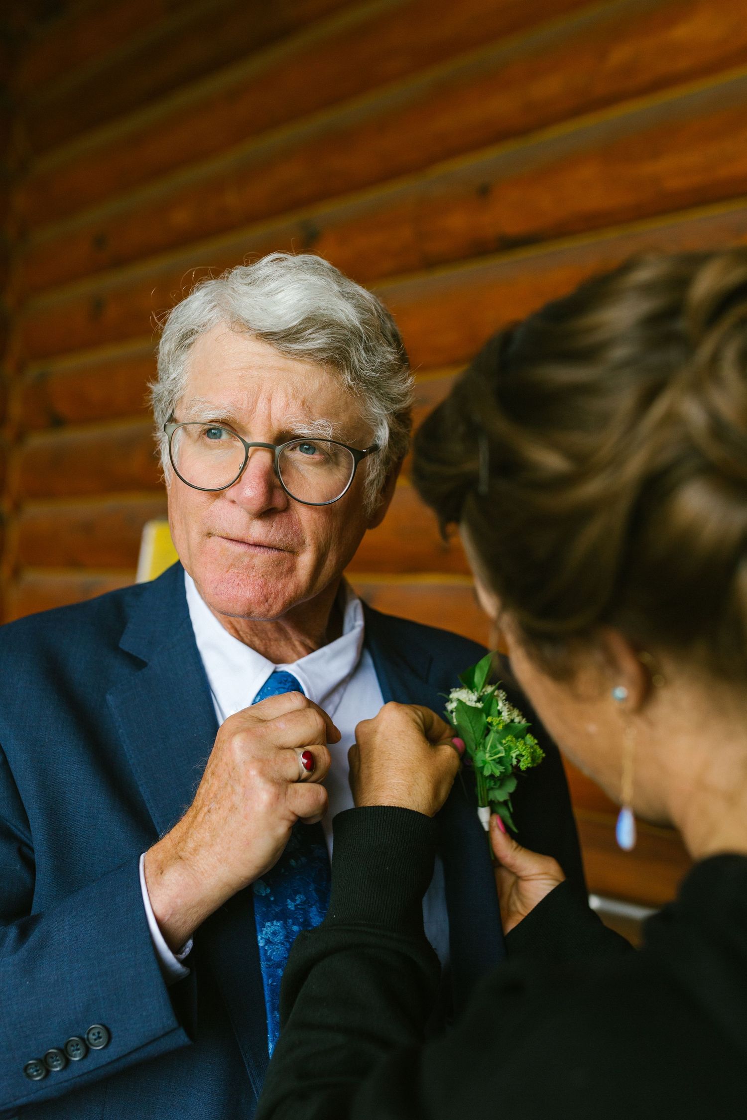 Someone wearing a navy blue suit and tie gets help with a green boutonniere from another person at a formal event.