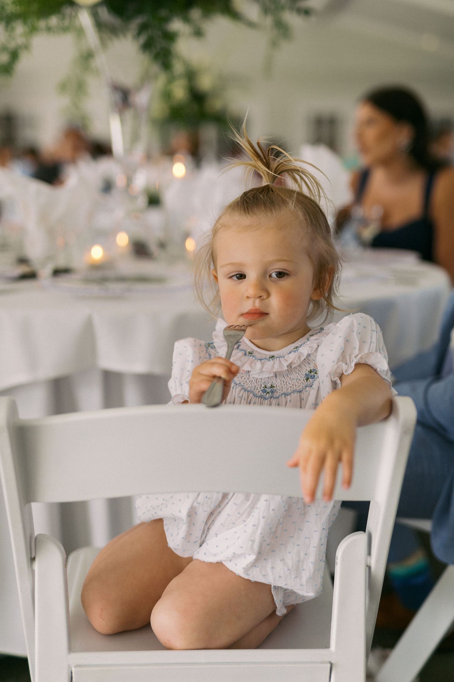 Small baby wearing white lace dress sits in white wooden chair at formal event with tables in background.