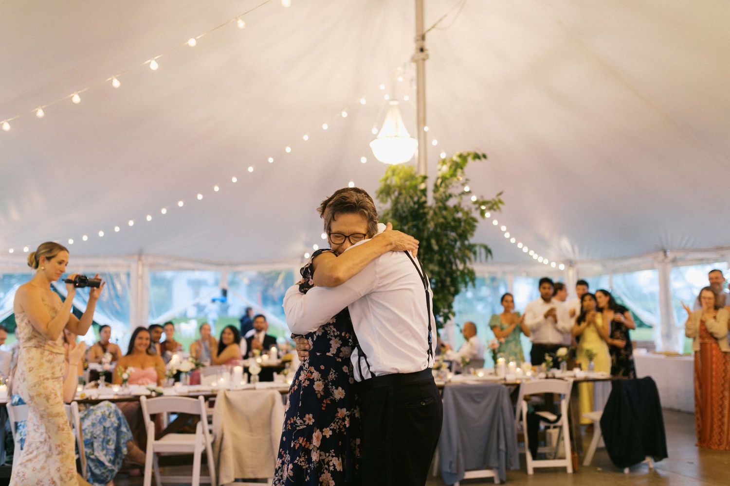 String lights illuminate an elegant white tent reception space with guests mingling and dining at round tables as a groom and his mother hug