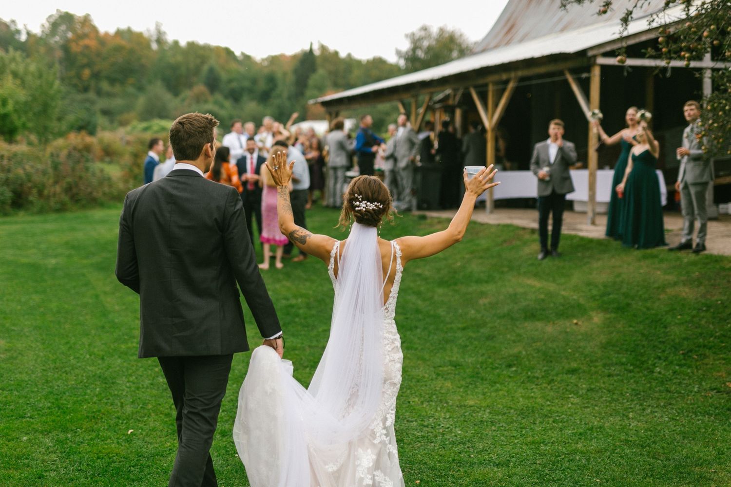 A couple in wedding attire raise their joined hands in celebration on a grassy lawn outside a rustic barn venue.