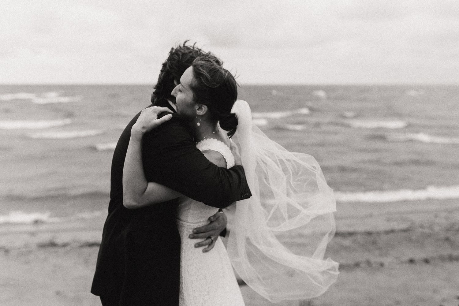 Black and white photograph of couple embracing on a windy beach with flowing veil.