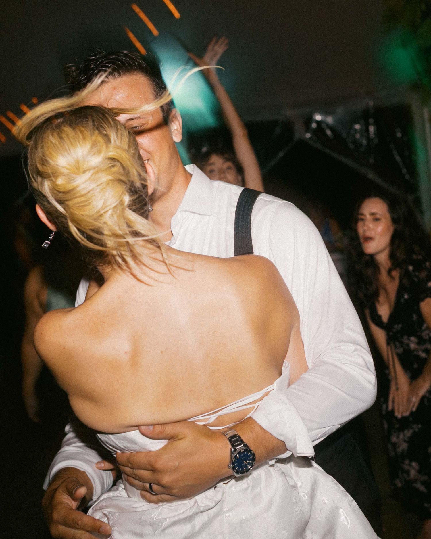 A just married bride and groom blur by the camera as they dance closely together at a dimly lit reception as guests cheer around them.