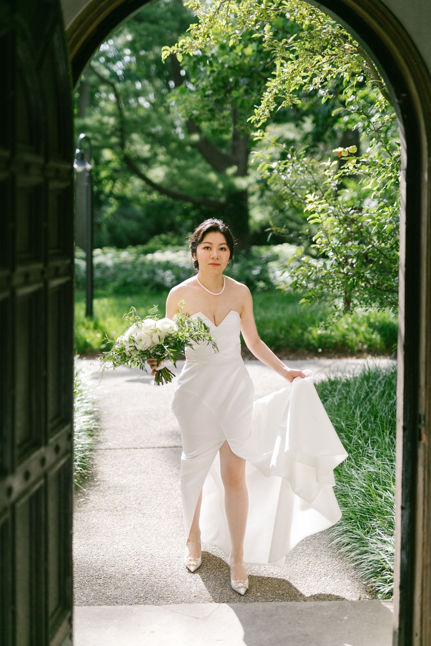 Bride in strapless white gown holds bouquet walking through arched doorway in lush green garden.