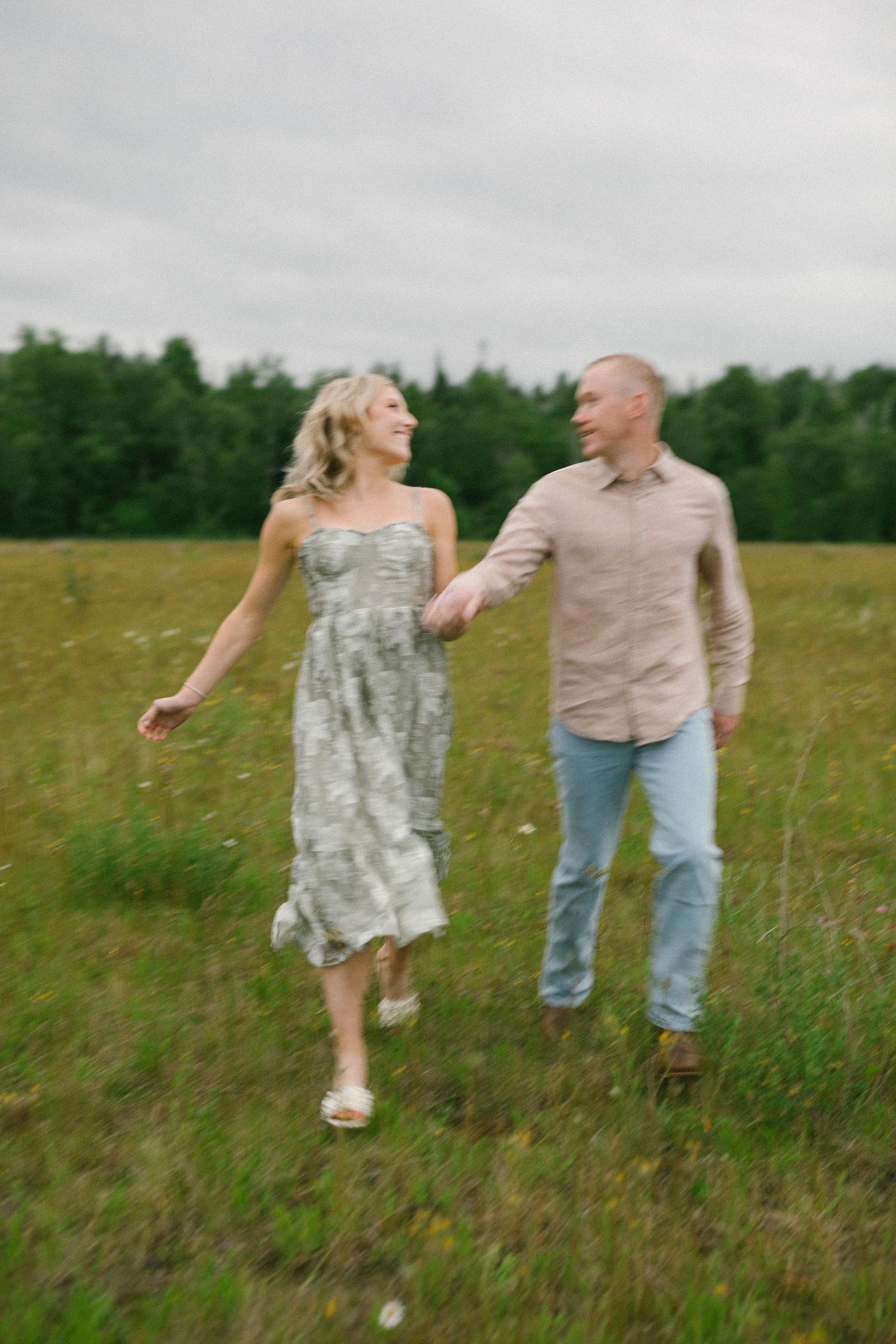 Joyful couple laughs together while walking through a summer meadow with grey skies overhead.