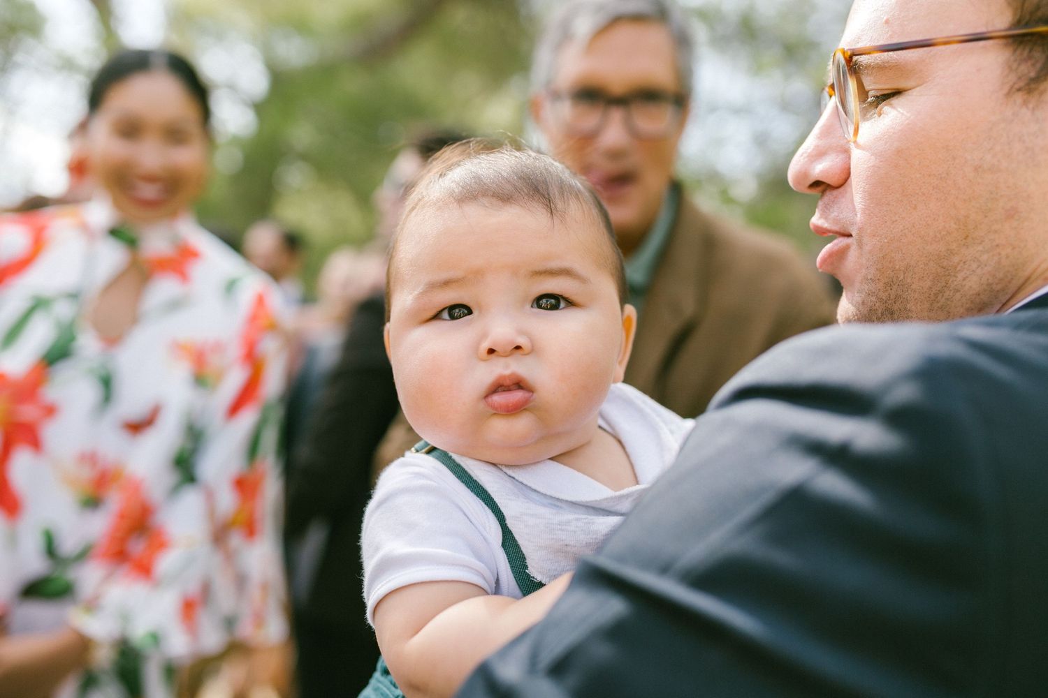 A baby wearing a white shirt is being held outdoors while two people look on in the background.