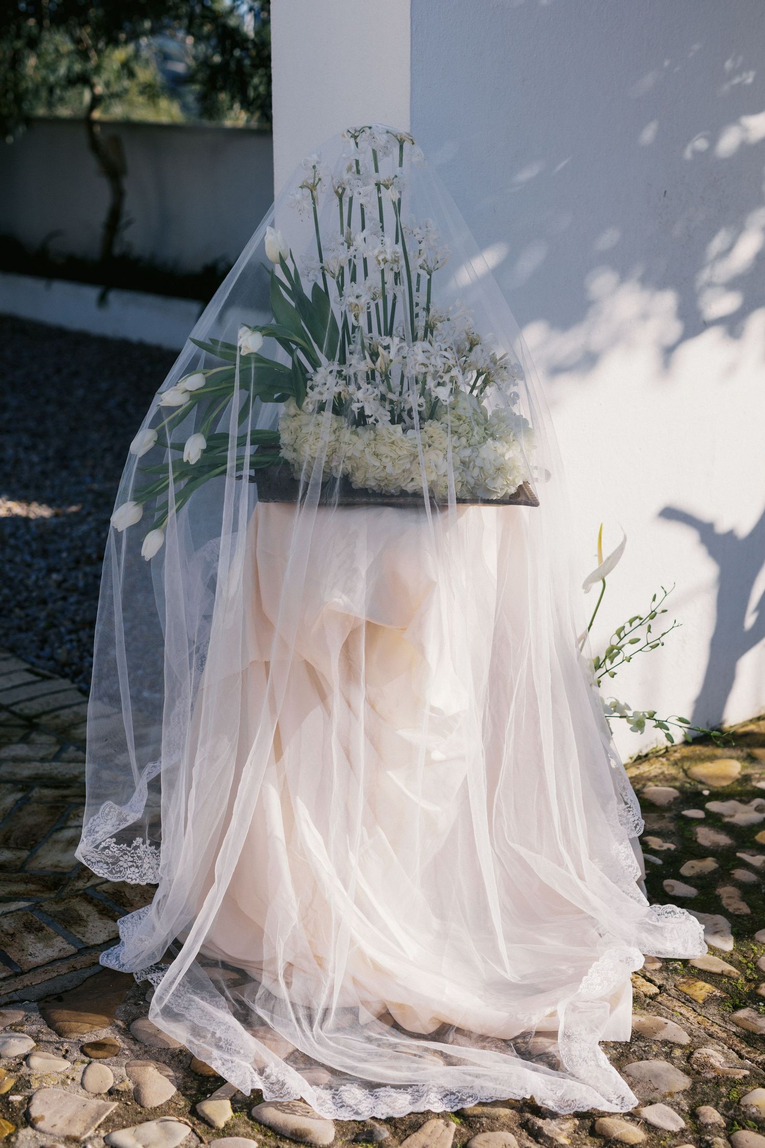 Bridal veil draped over floral arrangement on pedestal, with white tulips and delicate blooms in outdoor setting.