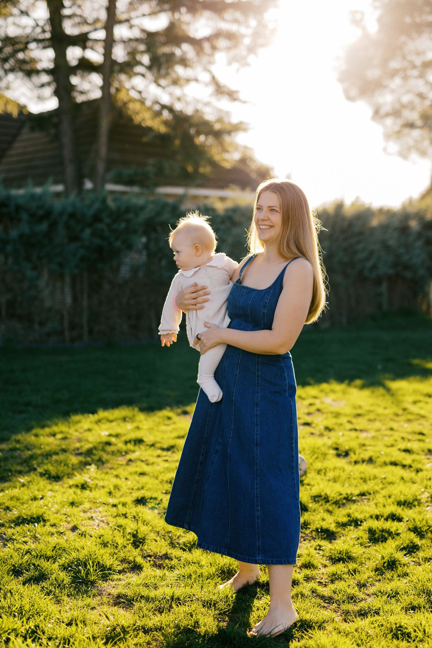 Smiling young mother in blue denim dress holding baby outdoors in sunny backyard garden.