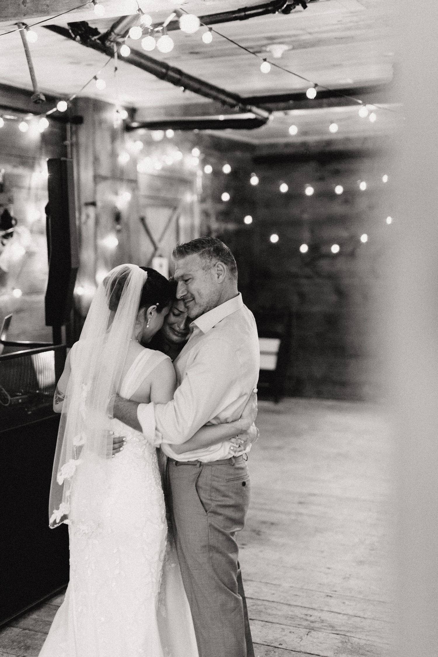 Black and white photo of two brides dancing with one father in a rustic barn with string lights glowing behind them.