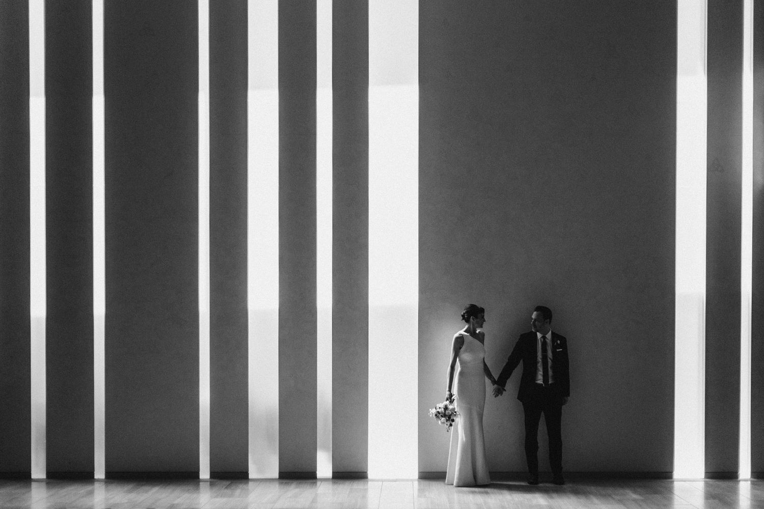 Black and white photo of bride and groom holding hands against striped light and shadow wall.