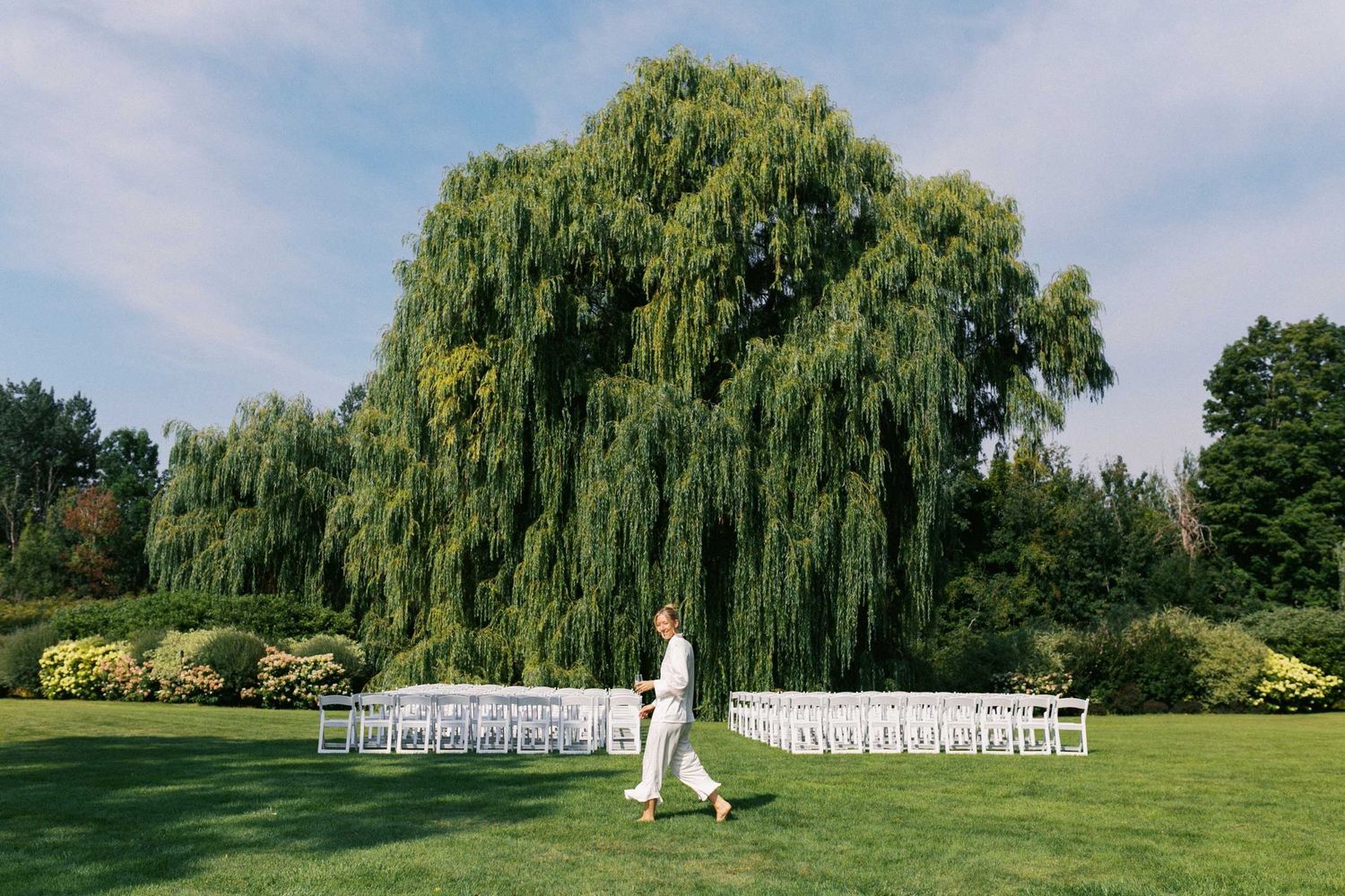 A bride walks in front of her ceremony scene of white chairs and a giant weeping willow tree on a lush green lawn.