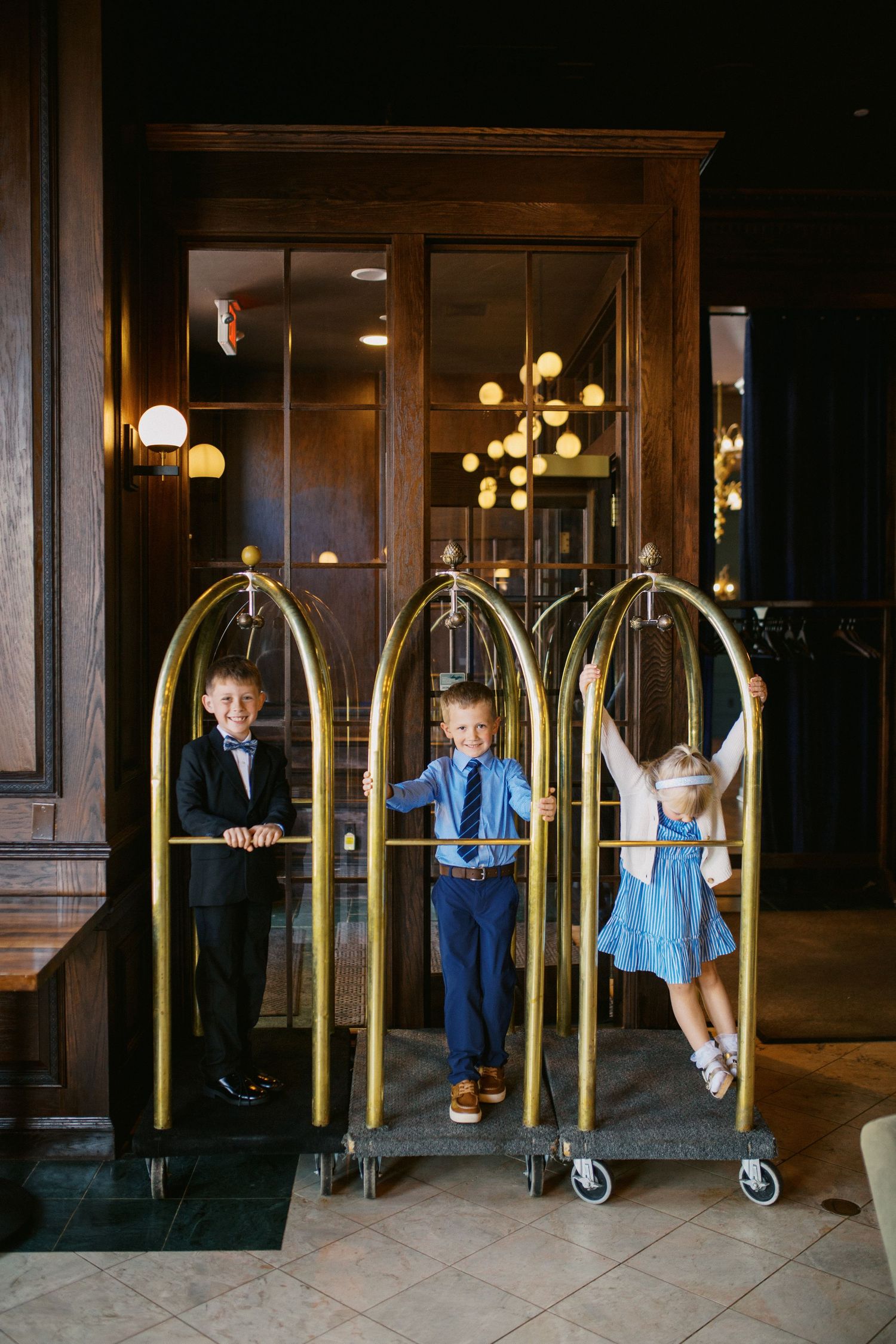 Three dressed-up children playing with golden hotel luggage carts in an elegant hotel lobby.
