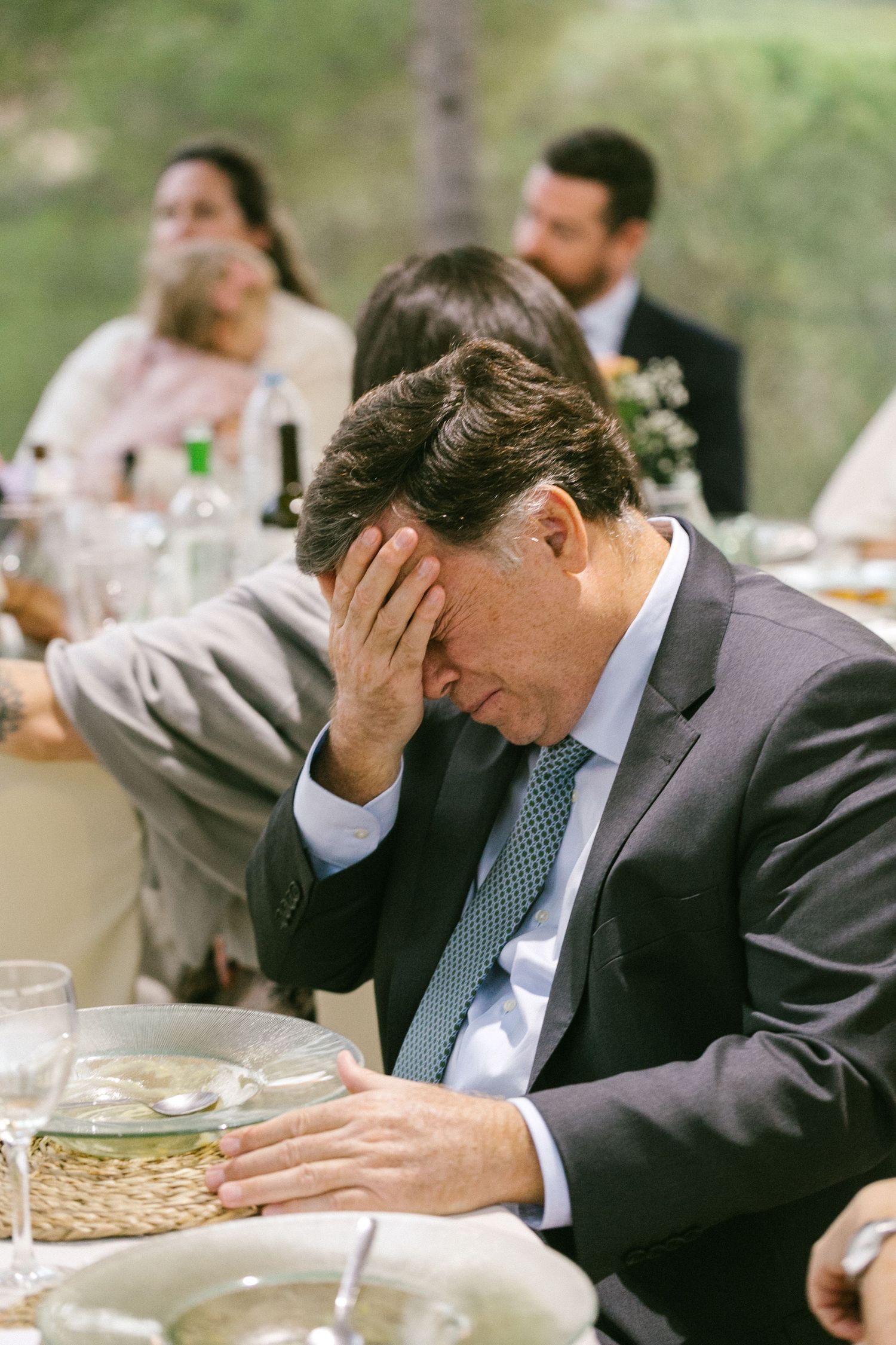 A guest in a business suit rests head in hand while sitting at an outdoor wedding reception with woven placemats.