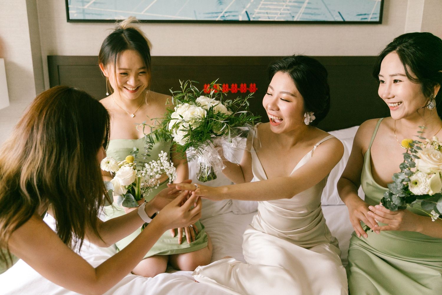Bride in white gown laughing with bridesmaids in sage green dresses while exchanging bouquets of white flowers on a bed.